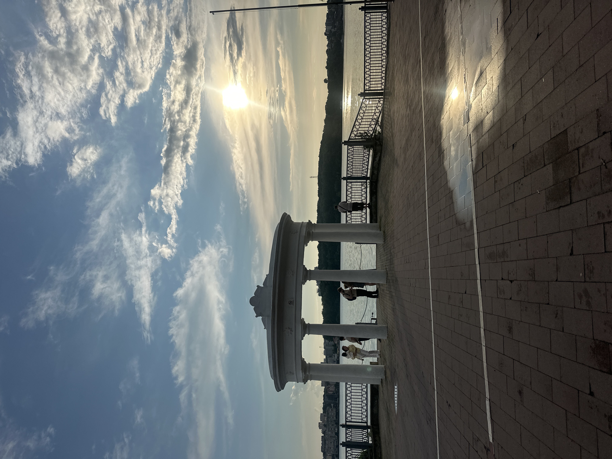 The image depicts a waterfront promenade in Ternopil, Ukraine, likely the Ternopil Pond embankment, given the presence of a large body of water, a city skyline in the distance, and a gazebo. The sun is setting, casting a warm glow and creating reflections on the wet paving stones in the foreground. Several people are visible: a group of three individuals stand under the gazebo, one of whom is holding a child. Another person stands near the railing, looking out at the water. A lone figure stands on the far right of the promenade, facing away from the camera. The architecture suggests a public park or recreational area, with decorative fencing lining the promenade. The sky is a mix of blue with scattered clouds, illuminated by the setting sun.