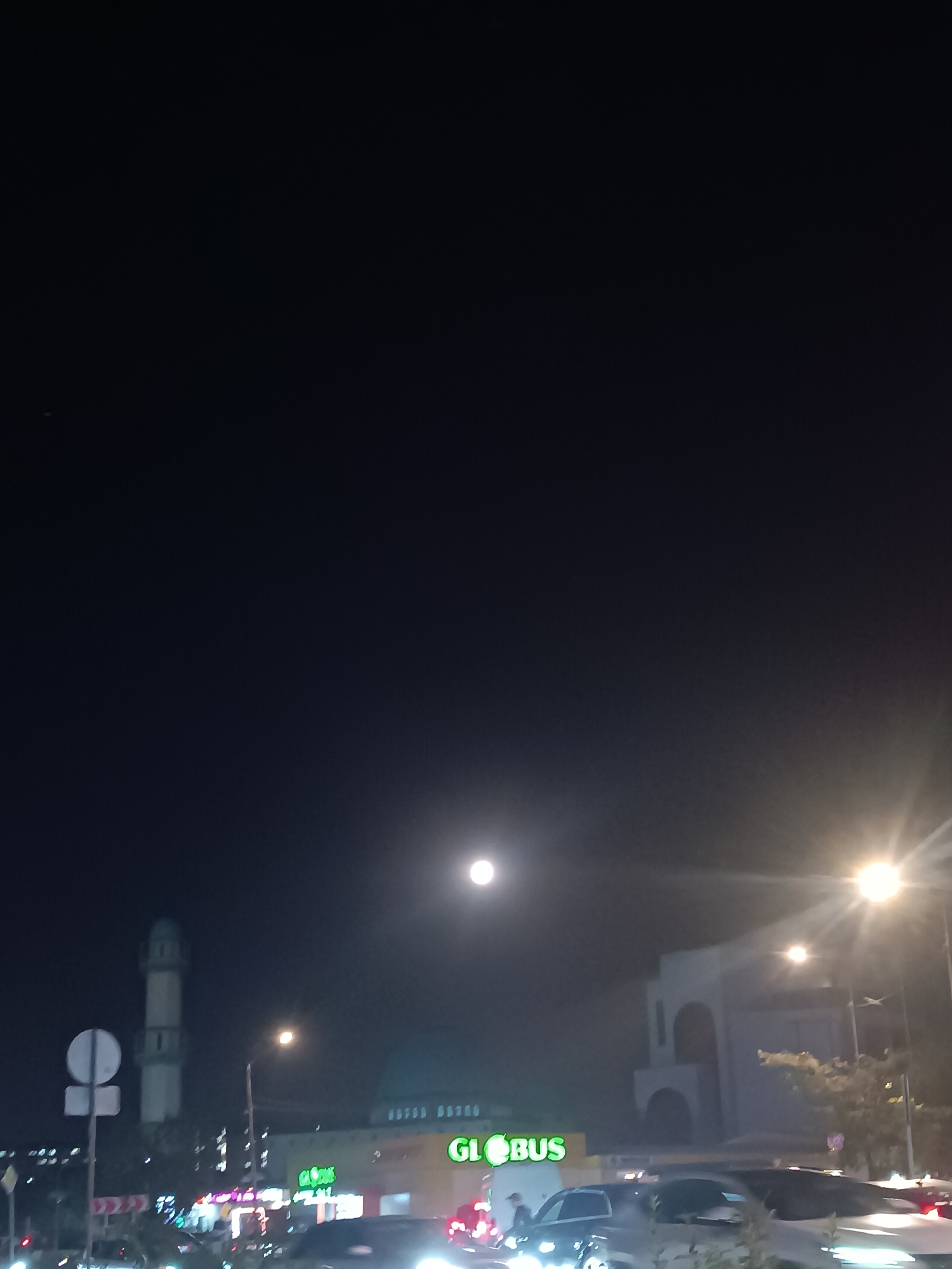 The image captures a night scene in Bishkek City, Kyrgyzstan, with a full moon prominently displayed in the sky. In the foreground, a street scene unfolds with several cars, some of which are moving, indicated by the headlights. A prominent "GLOBUS" supermarket sign is visible, illuminated in green, suggesting a commercial area. To the left of the supermarket, a minaret of a mosque stands tall, indicating the presence of a religious site. The lighting from street lamps and the moon creates a somewhat hazy, atmospheric effect. The overall impression is that of a typical urban night, with elements of commerce, religion, and transportation coexisting.