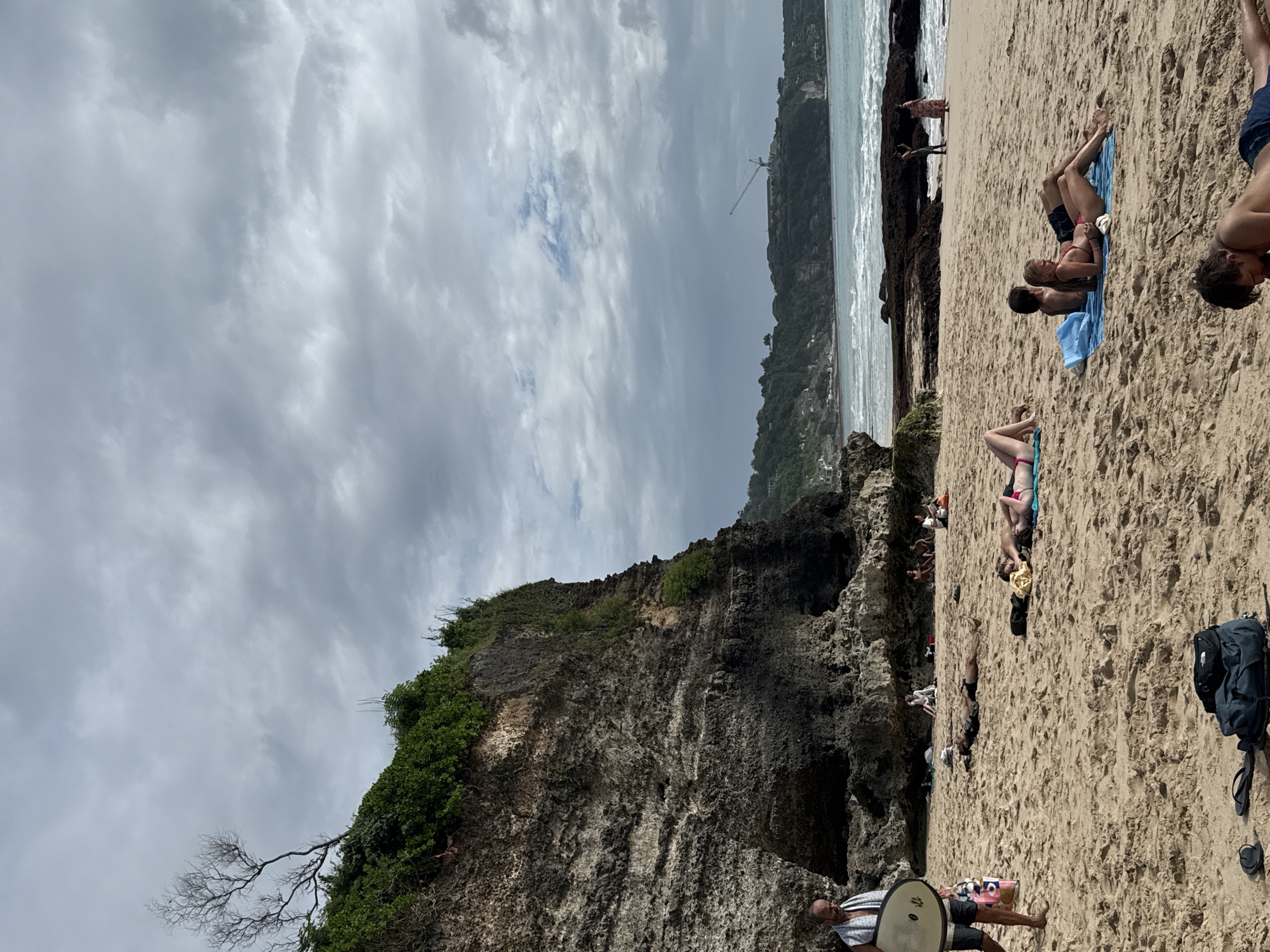 The image shows a beach scene in Kuta Selatan, Indonesia. A rocky cliff covered with green vegetation is visible on the left side of the image, with the ocean and sky in the background. The sky is overcast with grey clouds. On the beach, several people are sunbathing and relaxing on the sand. Some are lying on towels, while others are walking or standing near the water. A person carrying a circular object, possibly a surfboard or paddleboard, is walking near the bottom of the image. A backpack and sandals are visible on the sand on the lower right.