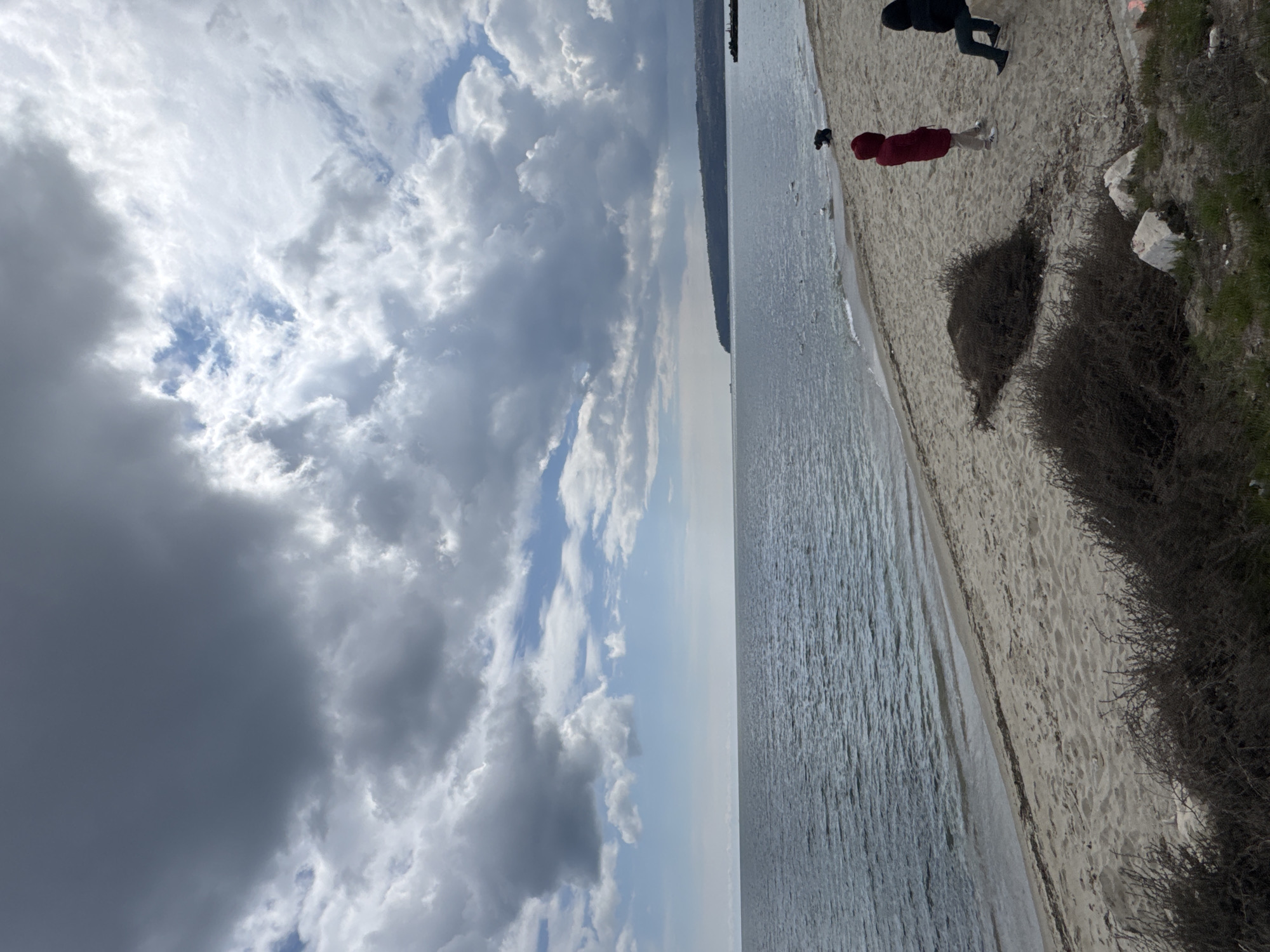The image depicts a view of the beach and the Black Sea in Varna, Bulgaria, under a partly cloudy sky. A narrow strip of land runs along the top right of the image, where the beach meets vegetation. Two people are visible on the beach. One is wearing a red jacket, and another is wearing a black jacket. The sea is calm. The left side of the image captures a dynamic sky with clouds in varying shades of gray and white, with patches of blue. The horizon line is visible in the center, where the sea meets the sky.