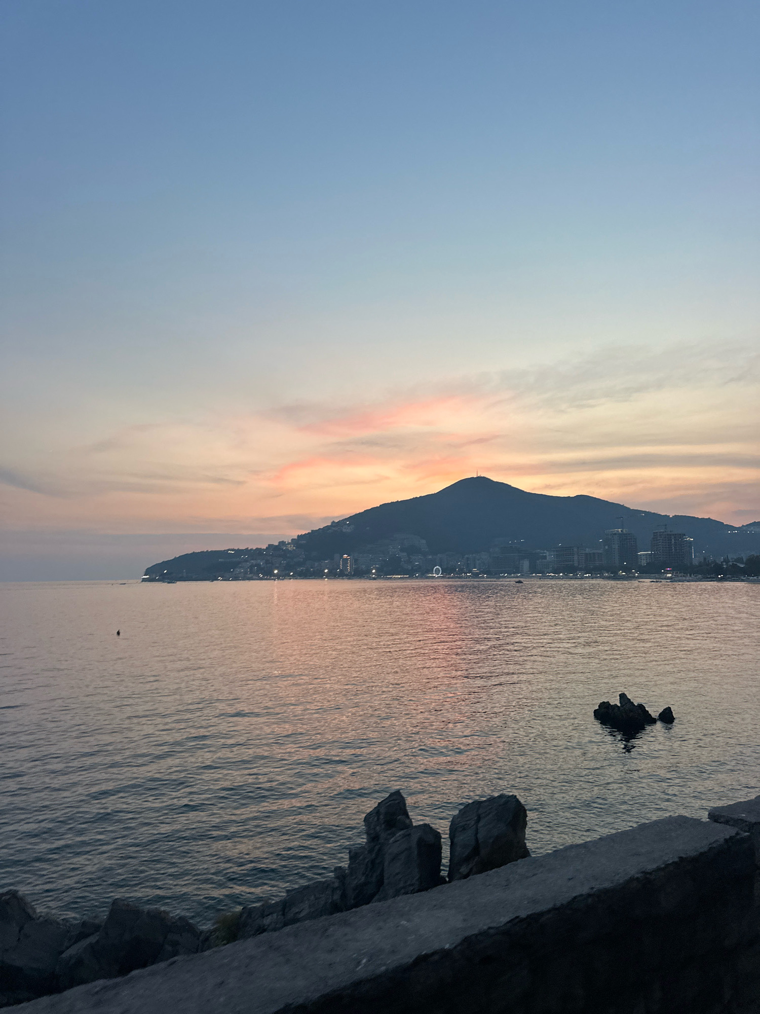 A twilight view of the Adriatic Sea coast in Budva, Montenegro, shows a calm, shimmering body of water reflecting the pastel hues of the sky. A silhouetted mountain range with a prominent peak, possibly the Ostrog Monastery on a far hill, dominates the background. The mountainside is dotted with the lights of buildings, suggesting a densely populated urban area. A Ferris wheel and other structures are visible on the coastline. In the foreground, large rocks are piled along a concrete embankment, with a few dark rocks protruding from the water. The sky transitions from a pale blue at the top to soft pinks and oranges near the horizon, indicating the sun has recently set. A small, dark buoy or marker floats in the water in the mid-ground. The overall impression is one of serene coastal beauty at dusk.