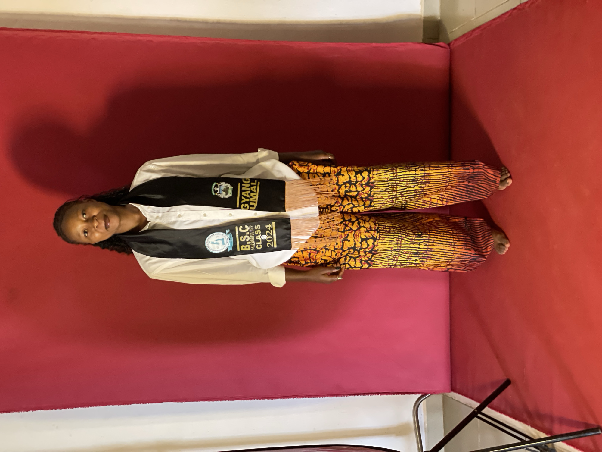 A full-shot, eye-level, indoor studio photograph features a young Black woman standing center frame against a vibrant red backdrop. The woman, wearing a white collared shirt and a black sash over it that reads "B.SC MICROBIOLOGY CLASS 2024" and "GYANG UMAL", is also adorned in loose-fitting, brightly patterned orange and yellow pants. Her dark hair is styled in braids that fall over her shoulders. She stands barefoot on a red mat, her hands by her sides. To the left of the frame, a black metal tripod with a light stand is partially visible. The lighting is direct, casting a shadow of the woman onto the backdrop. The overall setting suggests a celebratory or commemorative portrait, likely within a photography studio. There is no discernible indication of the specific location within Durumi, Nigeria.