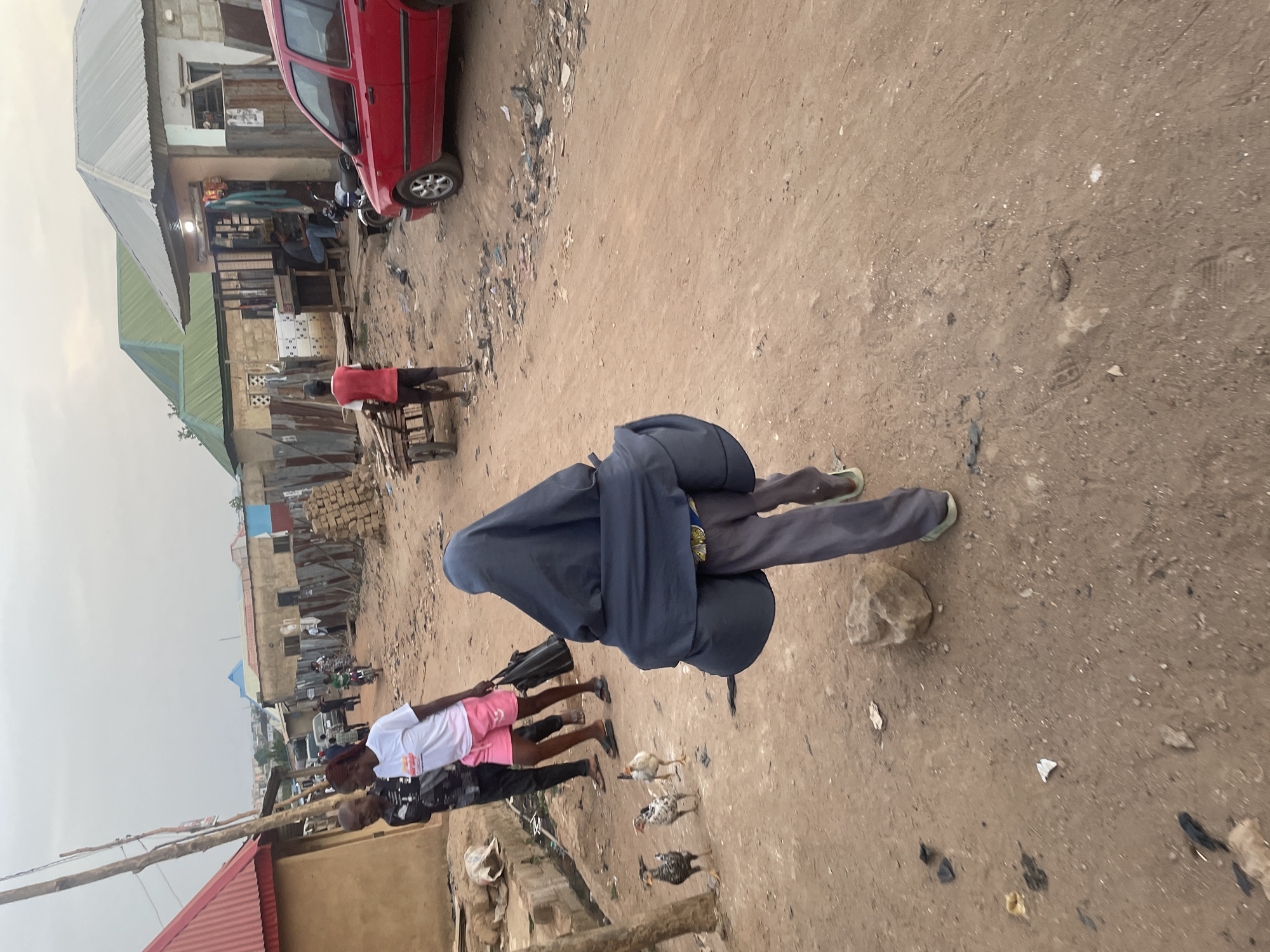 This image captures a bustling street scene in Abuja, Nigeria, characterized by an unpaved dirt road under an overcast sky. The ground is a sandy-brown earth, littered with small rocks, debris, and refuse, with visible tire tracks and footprints.
In the foreground, facing away from the camera, a person wears a dark blue full-body covering and trousers, with light-colored slippers visible. Near them, three chickens — two speckled black and white, one lighter brown — are seen pecking at the ground.
Further into the scene, several people are visible. Two individuals walk side-by-side, one wearing a white t-shirt and pink shorts, the other in dark attire. To the left, a person in a red shirt is near a wooden pushcart. In the mid-ground, a bright red compact car is parked on the dirt road, partially visible.
The left side of the frame is dominated by a series of simple, low-rise buildings with corrugated metal roofs, some grey, some green. These structures appear to be local shops or residences, with people gathered around or inside. One building shows a person seated, and a motorcycle is parked nearby. In the background, more buildings and piles of construction blocks suggest an ongoing development or a dense urban environment. The general activity indicates routine daily life in a lively community. No discernible text is visible.