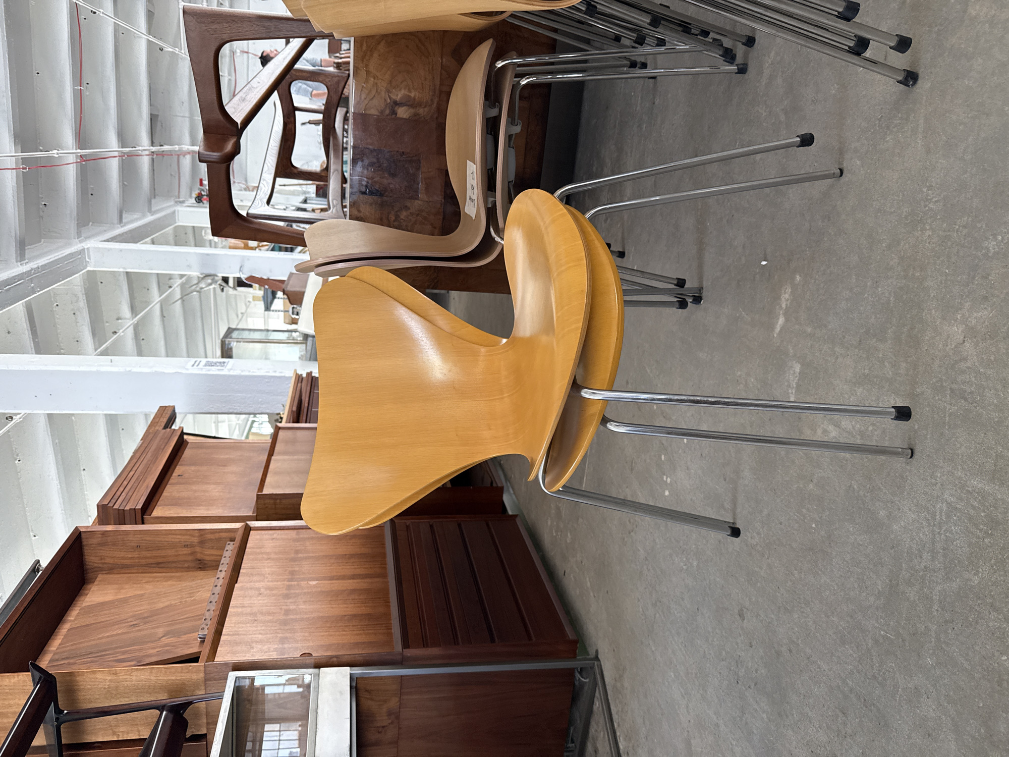 The image shows a collection of stacked chairs and wooden furniture, likely in a warehouse or storage facility. The chairs have a light wood finish with metal legs. Some of the wooden furniture pieces appear to be drawers or cabinet components. The location could potentially be in New York City, as specified, but there are no distinct indicators to confirm this within the image itself. The focus is on the arrangement of furniture rather than a specific activity or interaction. The environment is utilitarian, suggesting a space for storing or displaying furniture items.