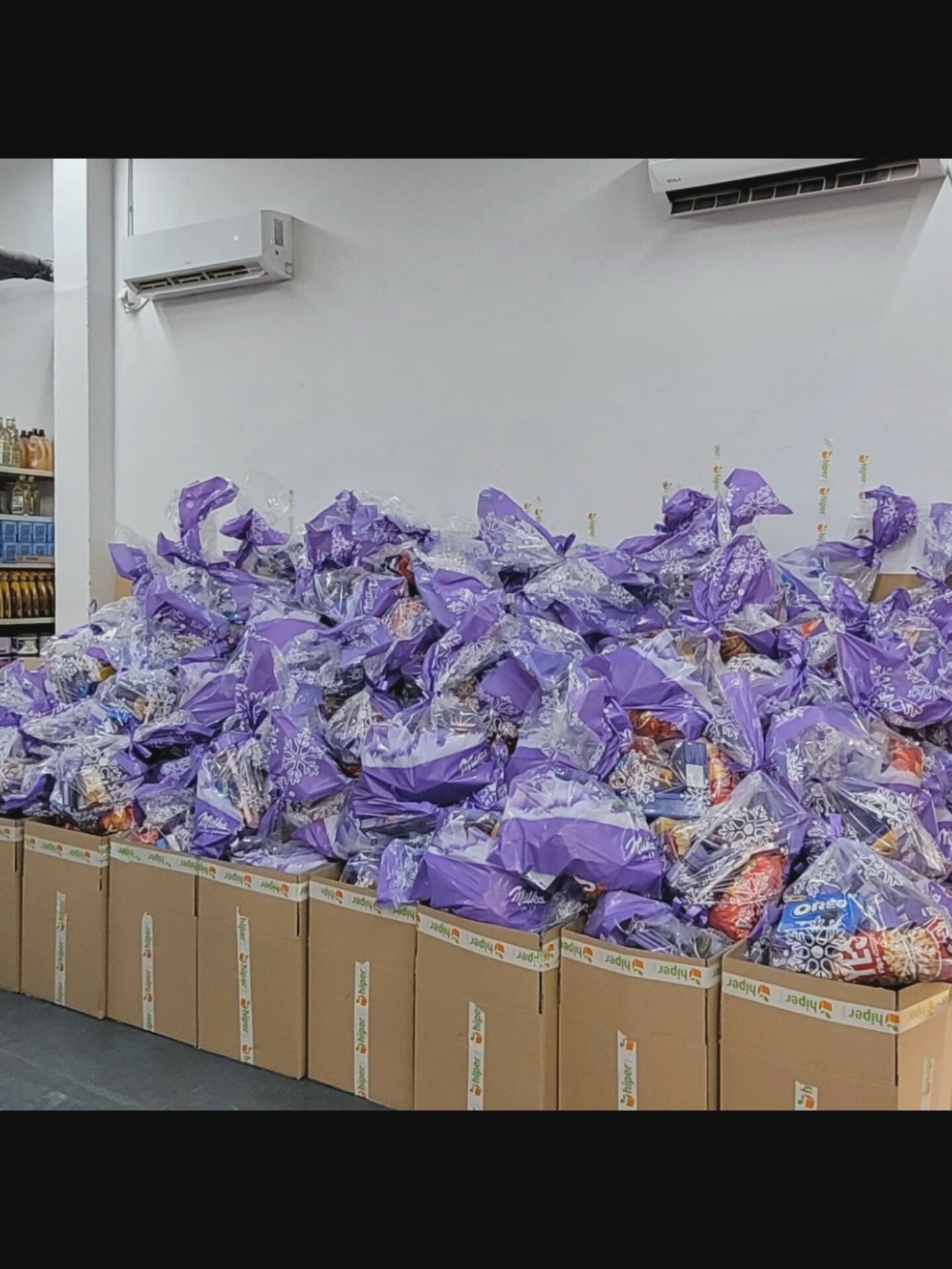 In Bielawska Wieś, Poland, a large pile of gift bags, predominantly purple and clear cellophane, is stacked on top of cardboard boxes. The gift bags are decorated with white snowflake patterns and tied with ribbon. Some bags appear to contain food items, with labels visible on some of the contents. The boxes are sealed with tape that has the "hiper" logo printed repeatedly. In the background, a white wall features two air conditioning units mounted high up. To the left of the gift bags, shelves are visible, stocked with bottles and containers. The scene appears to be indoors, possibly a storage area or a distribution point. There are no people visible in the image.