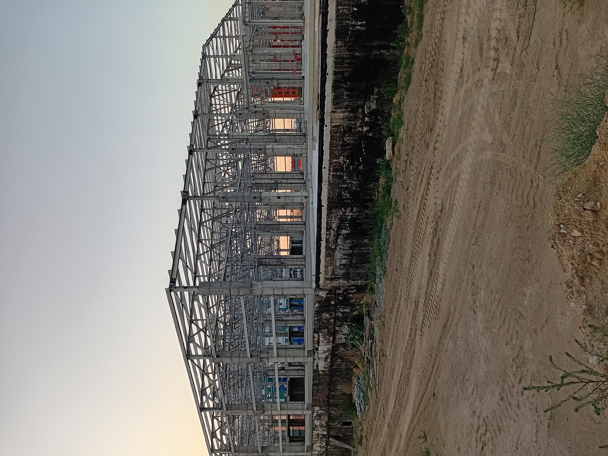 This is a medium-shot, eye-level, outdoor, vertical, color photograph of the skeletal frame of a large industrial building under construction at a site in Semey, Kazakhstan. The structure is made of grey metal beams and supports, with concrete pillars. The building appears to be a warehouse or a similar large-span structure, with an open roof and walls that are partially enclosed with what appear to be colorful modular units. The setting is a construction site with a dirt ground in the foreground, showing tire tracks from heavy machinery. The background features a clear, pale sky, suggesting either early morning or late evening light. There are no people visible in the photograph. The overall impression is one of ongoing development and construction.