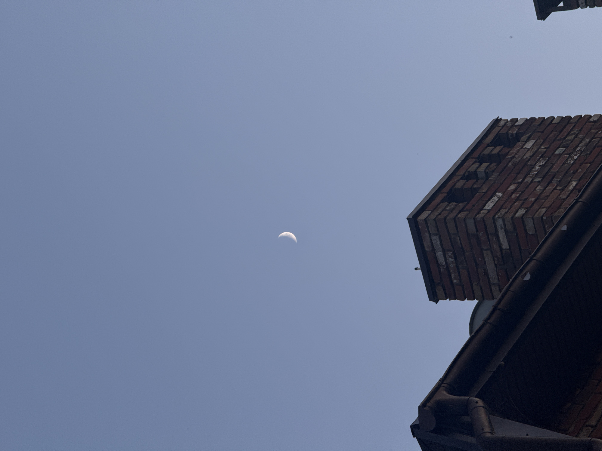A waning crescent moon is visible in a clear blue sky. In the foreground, the roofline of a building is visible, with a dark gutter and a brick chimney. The chimney is made of red brick and has decorative openings at the top. The scene appears to be an outdoor setting, possibly in the city of Obukhivka, Ukraine, given the context. No people are visible in the image.