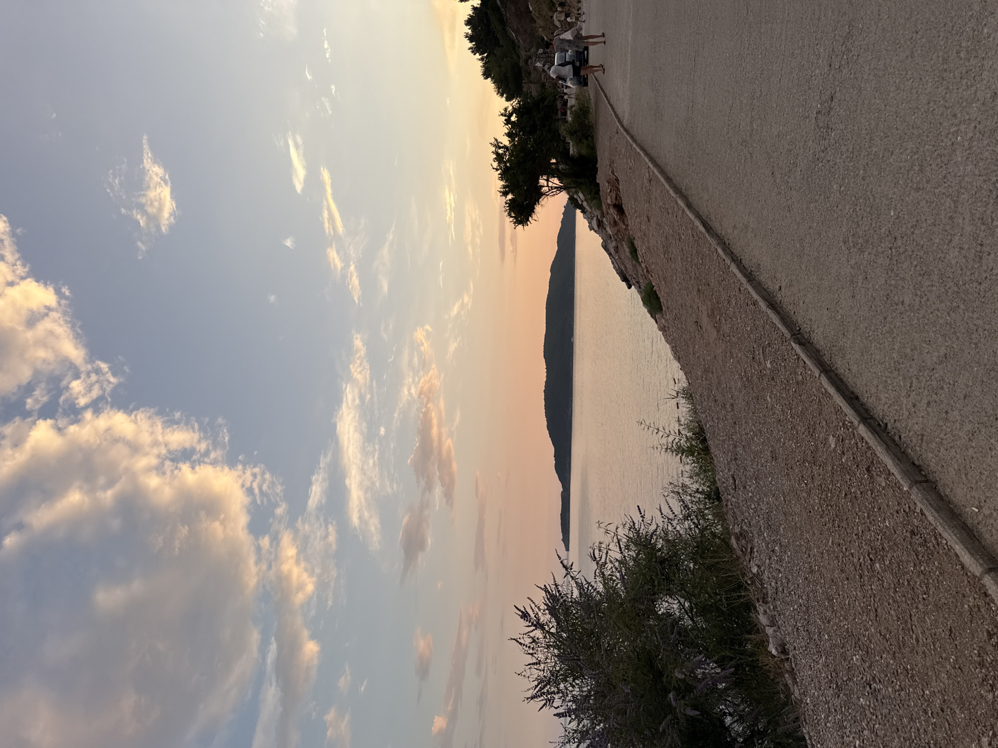 This media file captures a serene sunset scene at Kamenovo, Montenegro. The image features a picturesque view of a calm sea gently lapping against a coastline. In the foreground, a paved path runs alongside a low stone wall, with lush green trees and shrubs bordering the walkway.
Several people are visible on the path, appearing to be enjoying the sunset. They are casually dressed, suggesting a relaxed atmosphere. The sky is painted with soft hues of orange, pink, and blue, indicative of a sunset or dawn. Wispy clouds are scattered across the sky, catching the fading light. A distant, dark silhouette of a landmass, likely an island or coastline, is visible across the water.
The overall mood is peaceful and tranquil, evoking a sense of calm and appreciation for nature's beauty. The time of day is clearly sunset, given the warm, diffused light and the colors in the sky. The location is identifiable as Kamenovo, Montenegro. There is no visible text in the image. The scene is well-composed, balancing the natural elements of the sky and sea with the man-made path and wall.