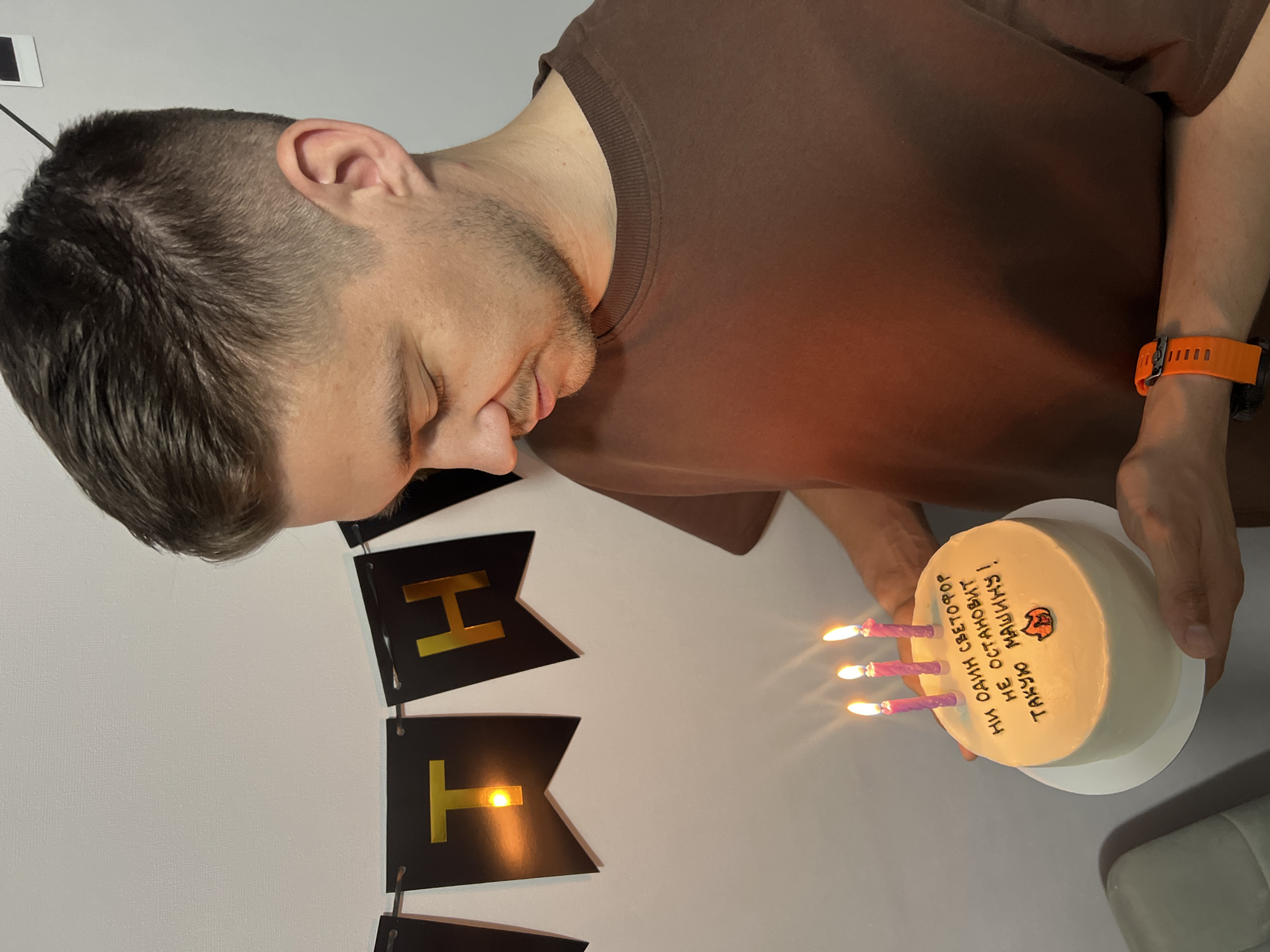 A man is celebrating his birthday. He is holding a small cake with three lit candles on it. The cake has a drawing of a tiger and a written message on it in Russian, which translates to "No traffic light can stop such a machine!". There is a black and gold birthday banner hanging behind him. The man has short, dark hair and is wearing a brown t-shirt and an orange smartwatch. The scene appears to be indoors, in a room with a white wall. The location is unknown, but the message on the cake suggests it might be Kazan, Russia.
