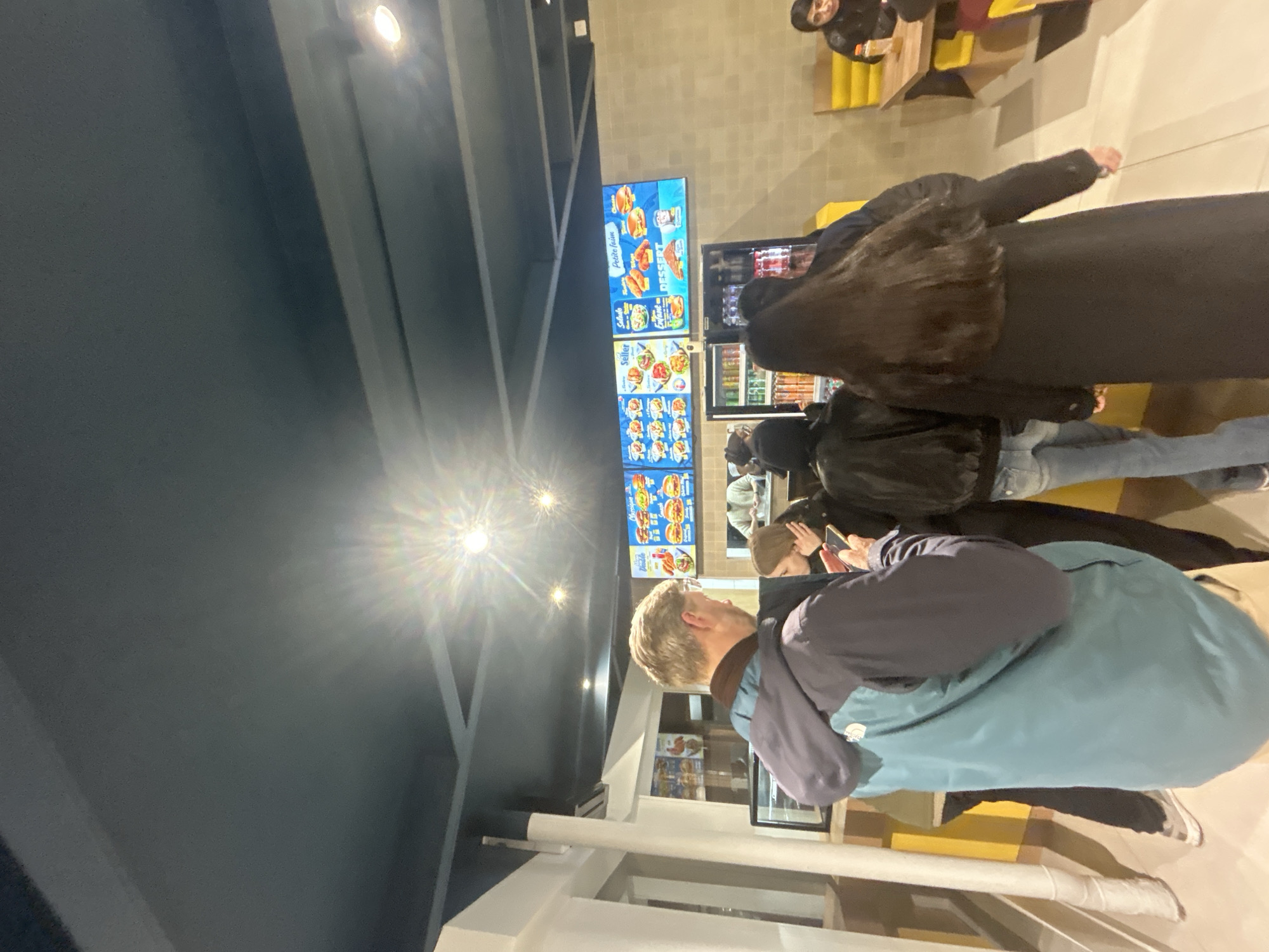 Here is a forensic description of the image:
Location: Noisy-le-Grand, France.
Scene: A group of individuals are standing in what appears to be a fast-food restaurant. Menu boards display images of food items above a counter. Customers are visible; some appear to be waiting to order. The architectural structure of the building has beams or supports. The artificial lighting seems to be bright and possibly LED.