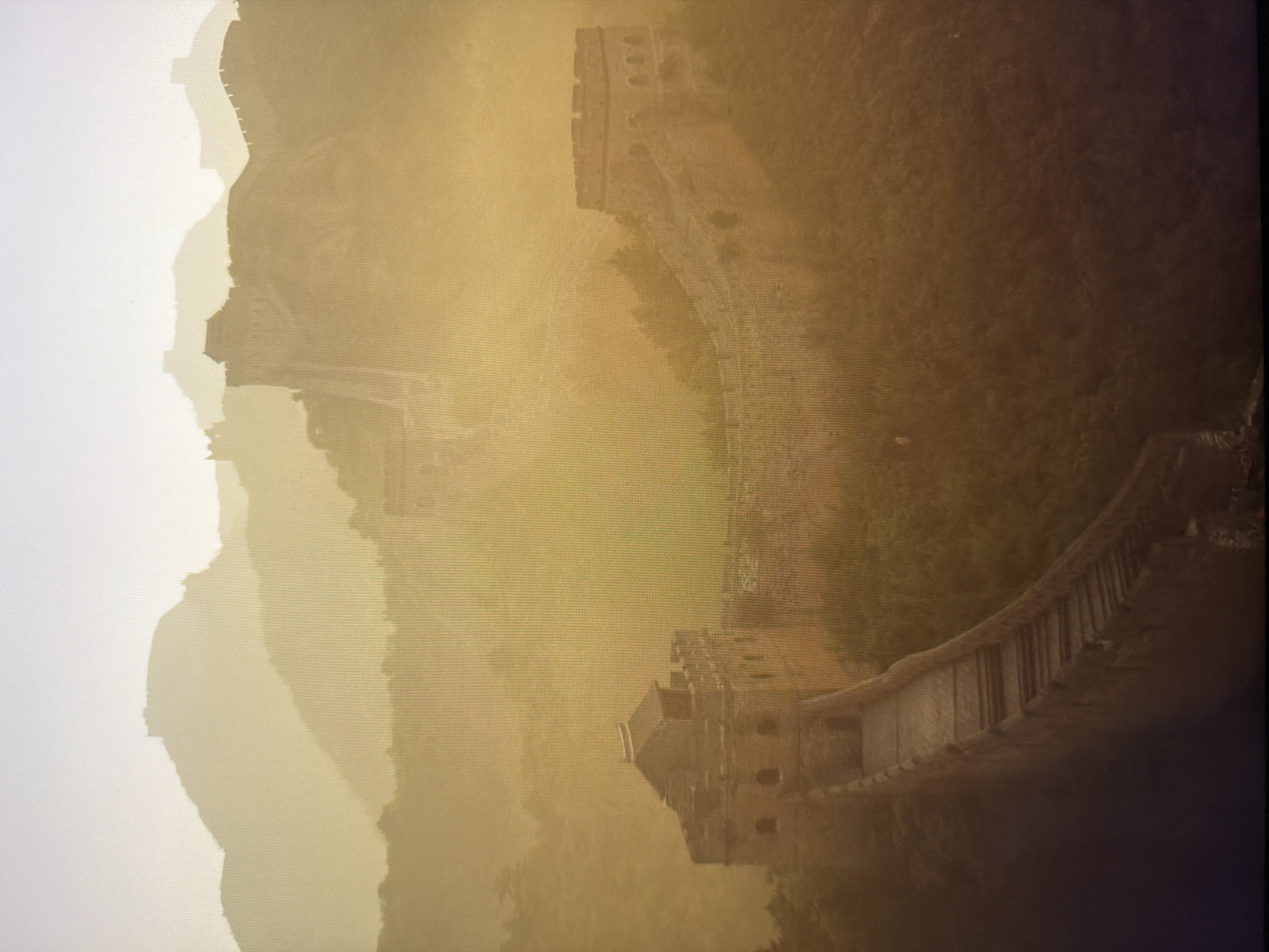 The image depicts a section of the Great Wall of China, characterized by its ancient stone construction winding over mountainous terrain. The scene is enveloped in a thick, golden haze, suggesting either a sunrise or sunset, or possibly dust or fog, which softens the distant mountain ranges and creates a layered, atmospheric effect. Multiple watchtowers and sections of the wall are visible, some in the foreground with greater detail, and others fading into the background. The wall itself appears to be made of brick and stone, with crenellations along the top. The surrounding environment is hilly and covered in dense vegetation, primarily trees and brush, which grow on the slopes. No people are present in the image. The overall impression is one of historical grandeur and natural beauty, muted by atmospheric conditions. The image does not depict any location in the city of Kyiv, Ukraine.