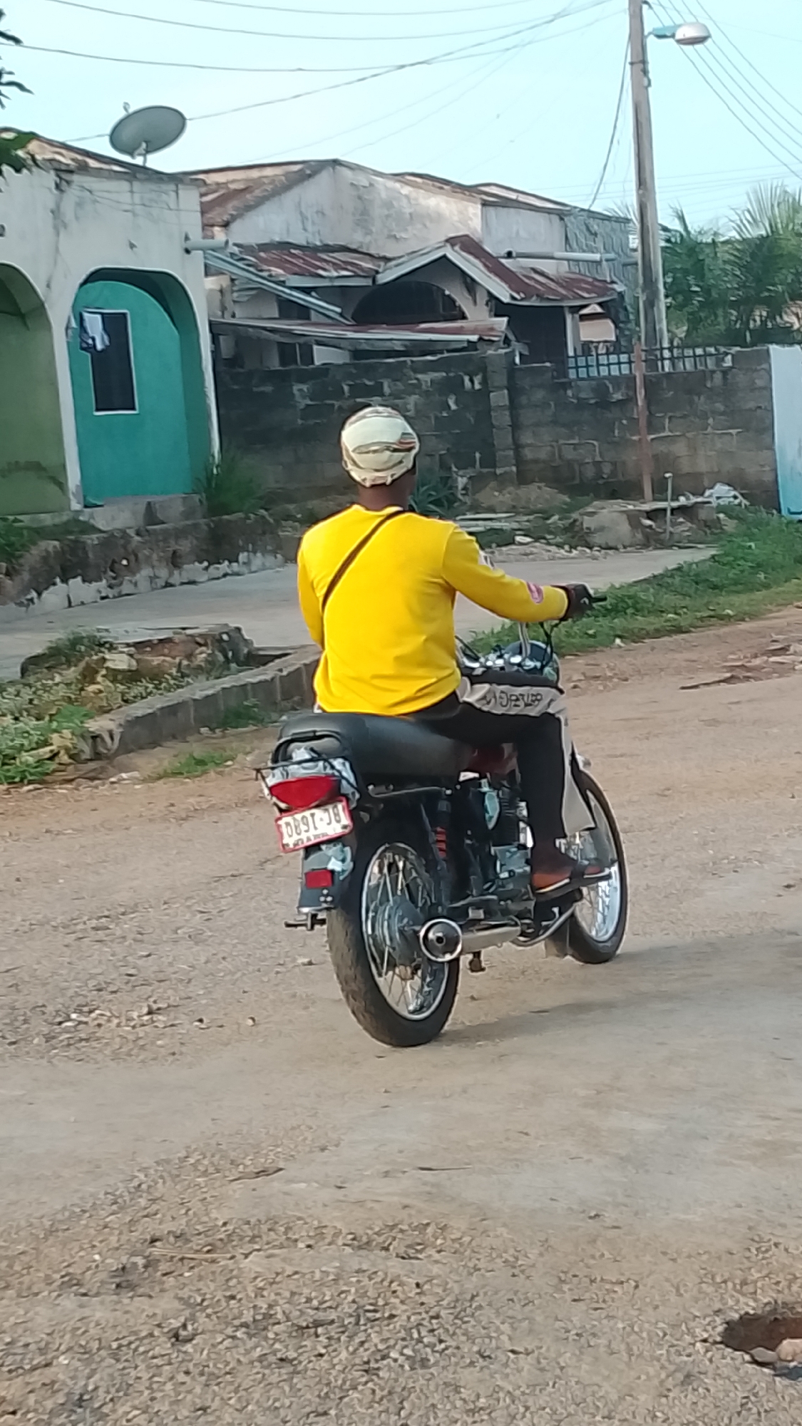 A motorcycle rider, viewed from behind, travels along an unpaved, gravelly road in what appears to be a residential or semi-urban setting. The rider wears a bright yellow long-sleeved shirt, dark trousers, and a light-colored helmet or head covering. A dark strap from a bag or satchel crosses their back.
The motorcycle is black and chrome, with a visible exhaust pipe and a rear license plate. The license plate clearly displays "BC-1680". Below this, smaller text reads "RC NO. A/WL" and "KWA", suggesting Kwara State, Nigeria. Partial text, possibly "SUPER", is visible on the motorcycle's tank or dashboard area.
In the background, several buildings are visible under a clear, bright sky. On the left, a light green and white building features an arched entrance and a satellite dish on its roof. Further back, other structures have discolored walls and corrugated metal roofs. A concrete block wall runs along the side of the road, and a utility pole with a street light stands on the right, with power lines crisscrossing above. The overall scene suggests a typical streetscape in Lokoja, Nigeria, consistent with the provided location context. The lighting indicates either early morning or late afternoon.