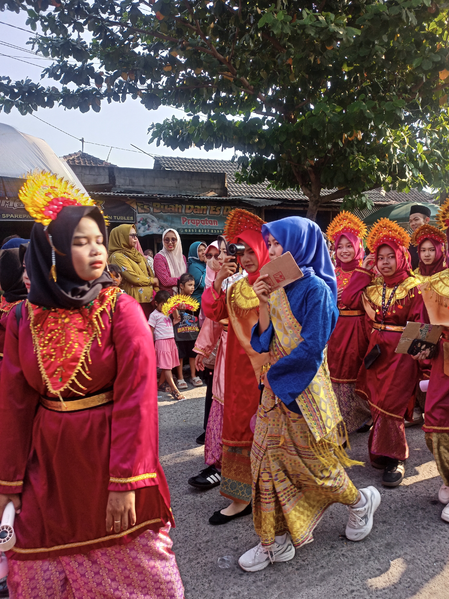 This is an outdoor, eye-level, full-shot photograph of a procession in Saren, Indonesia. In the foreground, a young woman wearing a traditional dark blue hijab and a brightly colored, patterned skirt is walking to the right. She is holding a small, beige-colored card in her right hand. To her left and slightly behind her, another young woman in a red traditional outfit and black hijab is walking, holding a small camera. Several other young women in similar red and gold traditional attire are visible in the middle ground, some wearing ornate headpieces. A group of younger children, some holding fan-like objects, stand behind them. A man wearing a black cap is visible in the background. The procession is taking place on a paved street with buildings and trees lining the background. The lighting suggests it is daytime, possibly in the late afternoon given the shadows.