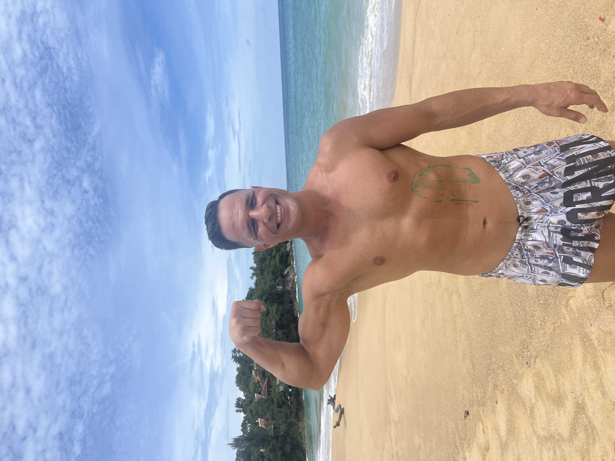 In Karon, Thailand, a man stands on a sandy beach, flexing his bicep for the camera. He has short, dark hair and is smiling broadly. He is wearing swim trunks with a pattern of US dollar bills. A green tattoo of a helmeted figure is visible on his abdomen. The ocean stretches out behind him, with gentle waves lapping at the shore. Lush green trees and buildings are visible on a hillside in the background. The sky is blue with scattered white clouds.