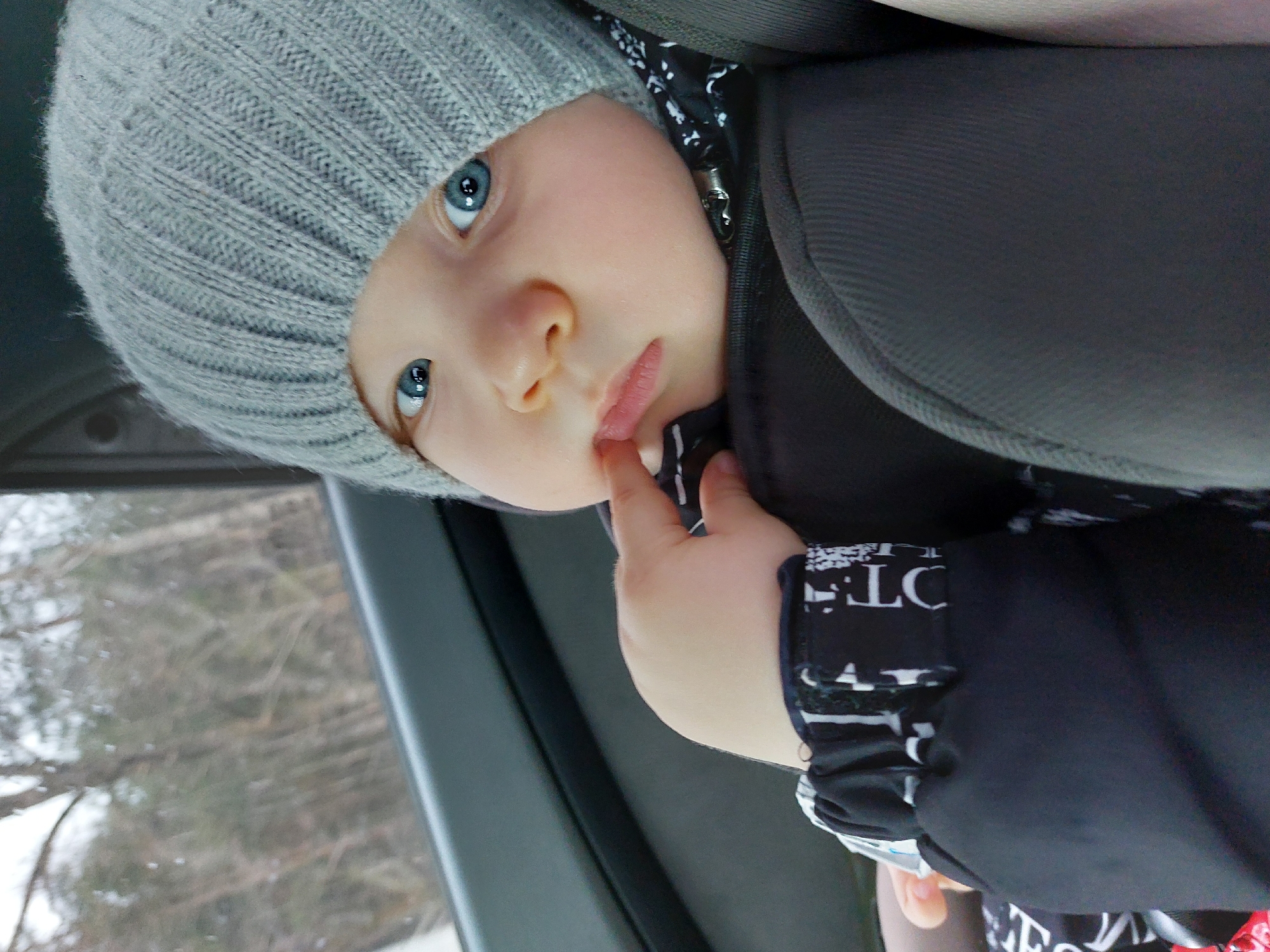 A young child with blue eyes and a gray knitted hat sits in a car seat. The child is wearing a black and white patterned garment and is touching their lips with their fingers. The car window shows an overcast, wintery scene, possibly with bare trees outside. Given the prompt, the location is assumed to be in or near Chaikovsky, Russia. The image appears to have been rotated counter-clockwise.