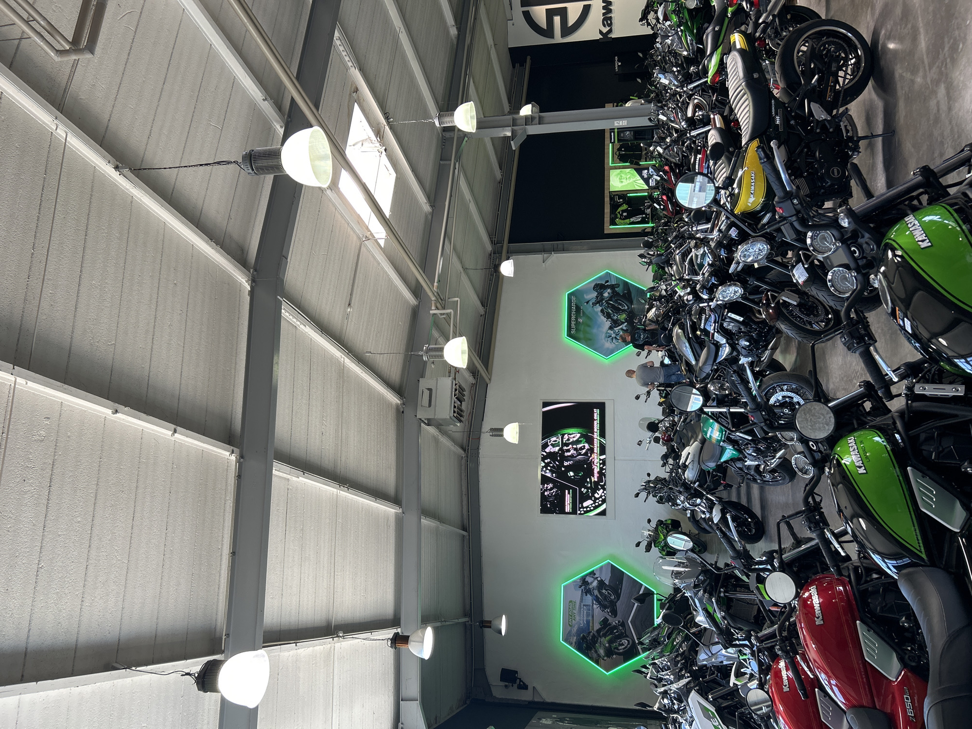 Here's a detailed description of the image:
**Setting:**
The image appears to be taken inside a Kawasaki motorcycle dealership or showroom. The interior is spacious with a high ceiling featuring exposed steel beams and white panels. Industrial-style lighting fixtures hang from the ceiling.
**Objects:**
* **Motorcycles:** A large number of Kawasaki motorcycles are displayed, primarily on the right side of the frame. Various models and colors are visible, including green, red, and yellow bikes.
* **Signage:** The Kawasaki logo is prominent, along with branding and marketing materials featuring the word "KAWASAKI", the text "LIMBACHER" and other textual information.
* **Wall Displays:** There are wall-mounted video display units showing animated speedometers and video content.
* **Neon Sign:** Some marketing displays feature a hexagonal shape with a green neon light outline.
**Subjects:**
* The main subjects are the Kawasaki motorcycles, showcasing the models available at the dealership.
**Actions:**
* The photo is a static shot intended to showcase the inventory and store layout.
**Environment:**
The overall environment is clean and well-lit, typical of a retail space. The motorcycles are neatly arranged to allow customers to view and potentially purchase them.