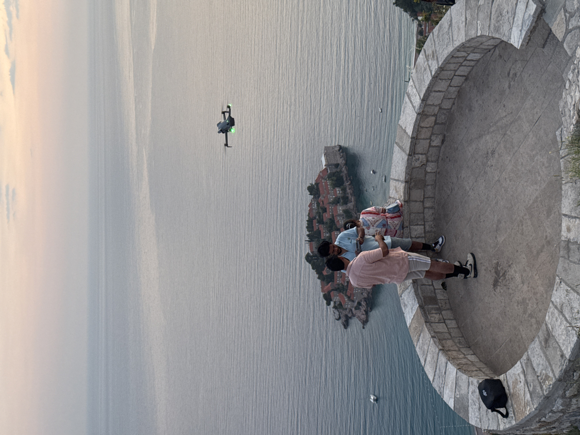 Two men are standing on a stone patio overlooking Sveti Stefan, Montenegro. One man wears a light blue shirt and the other wears a salmon-colored shirt. A drone is flying in the air near them and the island. The Adriatic sea is in the background, and a black backpack is on the ground near the patio wall.