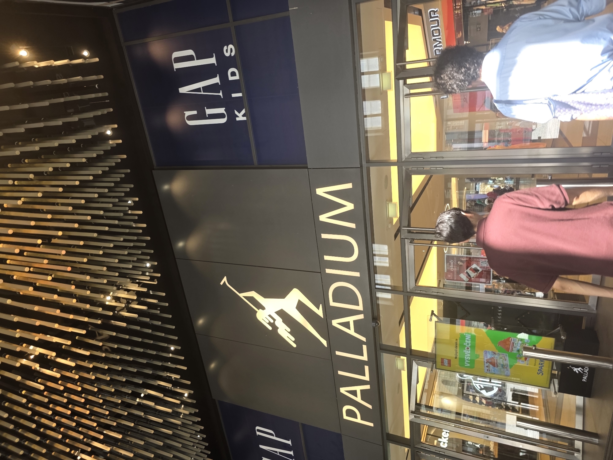 The image shows the entrance to a shopping mall with prominent signage for "PALLADIUM" and "GAP KIDS". The entrance features large glass doors and windows, with several people walking in and out. Inside, the mall appears to be brightly lit with yellow lighting. A large yellow advertisement with images of Lego sets is displayed in one of the windows. The ceiling of the mall's entrance area is adorned with a decorative installation of numerous thin, light-colored rods. The overall impression is that of a modern and busy retail environment.