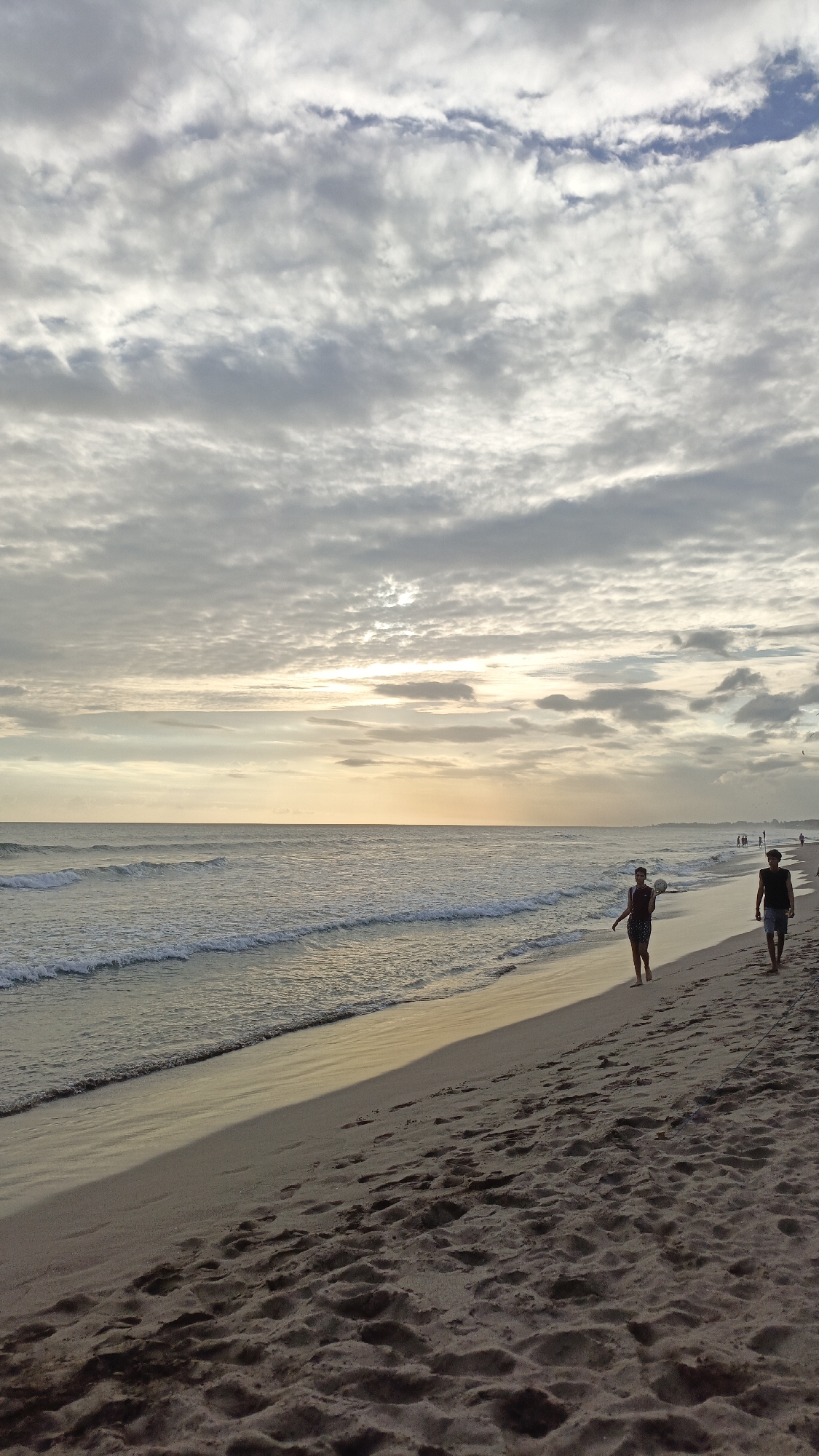 The image shows a beach scene in North Kuta, Indonesia, at sunset. The sky is filled with grey and white clouds, partially obscuring the sun. The ocean has gentle waves. Two people are walking along the sandy shore. One person is holding a ball, and the other is walking next to them. The beach is sandy with footprints.