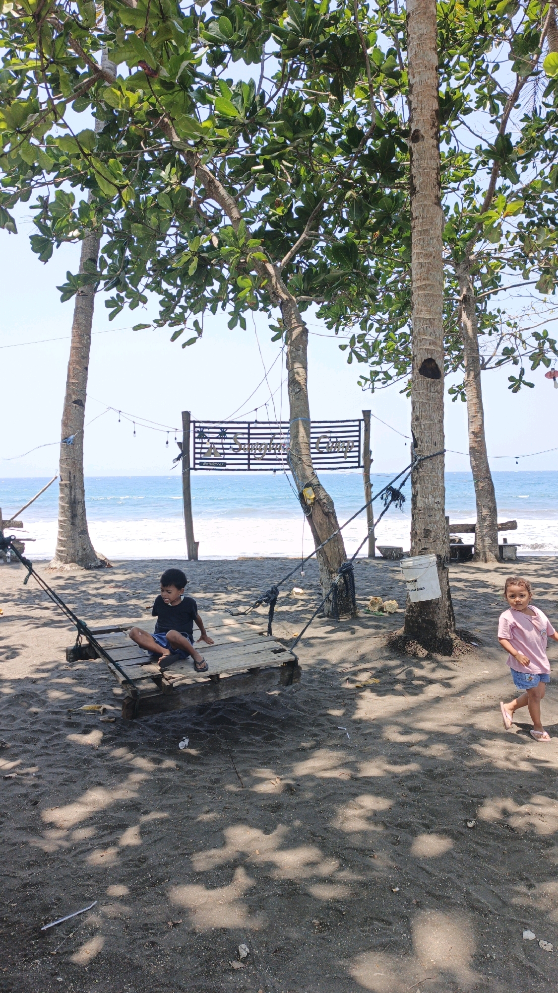 In Desa Batukaras, Indonesia, two children are at the beach, with palm trees and foliage overhead casting shadows on the sand. The ocean is visible in the background, with gentle waves lapping at the shore. A rustic wooden swing is set up on the sand, with one child, a boy in a black t-shirt and blue shorts, sitting on it, facing left. To the right, a young girl in a pink t-shirt and blue shorts is walking away from the camera, looking back. A sign with the words "A Sunghai Camp" and "Bira Karas" is visible between the trees.