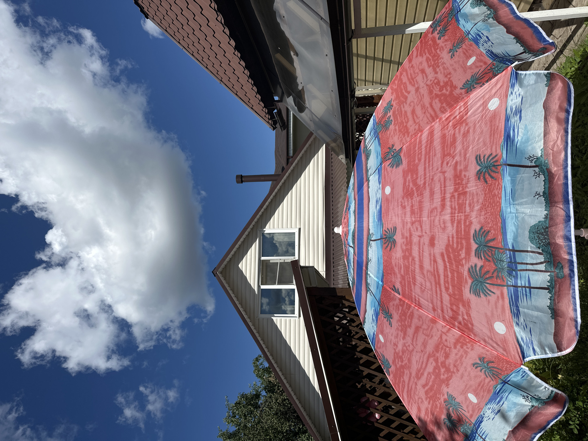 The image shows an outdoor scene with a house and a beach-themed umbrella. The house has a brown-tiled roof and light-colored siding, with two windows visible on the upper story. A chimney pipe protrudes from the side of the house. The umbrella is large and predominantly red, adorned with a tropical beach scene featuring palm trees, water, and moons. It is partially unfurled and appears to be the main focus of the foreground.
The setting is outdoors, under a bright blue sky with scattered white clouds. The sun is high, suggesting it is daytime. The presence of the umbrella hints at a sunny day, possibly for relaxation or outdoor activities. A wooden lattice structure is visible below the umbrella. Trees are also present in the background.
No people are visible in the image. The overall mood is bright and cheerful, evoked by the sunny weather and the colorful umbrella. There is no visible text in the image. The location context is Nizhny Novgorod, Russia.