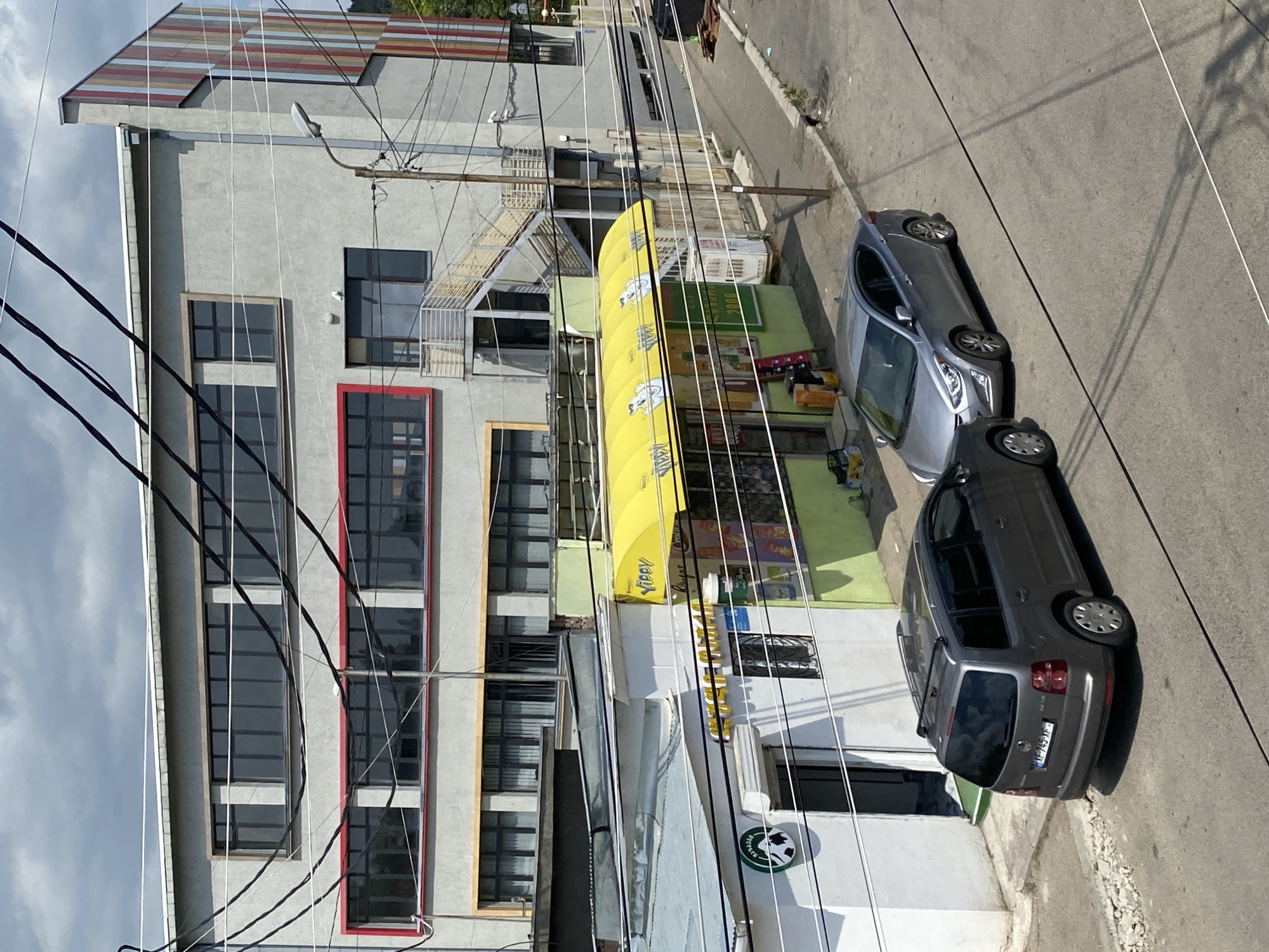This is a street-level, eye-level photograph taken on a cloudy day, looking toward a multi-story building with a light gray facade and a multi-colored vertical striped design on its right side. The building has several horizontal bands of windows. The lower portion of the image shows a row of shops with a bright yellow awning above one of them. A gray minivan is parked on the asphalt street to the left, and a silver sedan is parked to the right. Overhead power lines crisscross the scene, casting shadows on the buildings and street. The scene is likely in Tbilisi, Georgia.