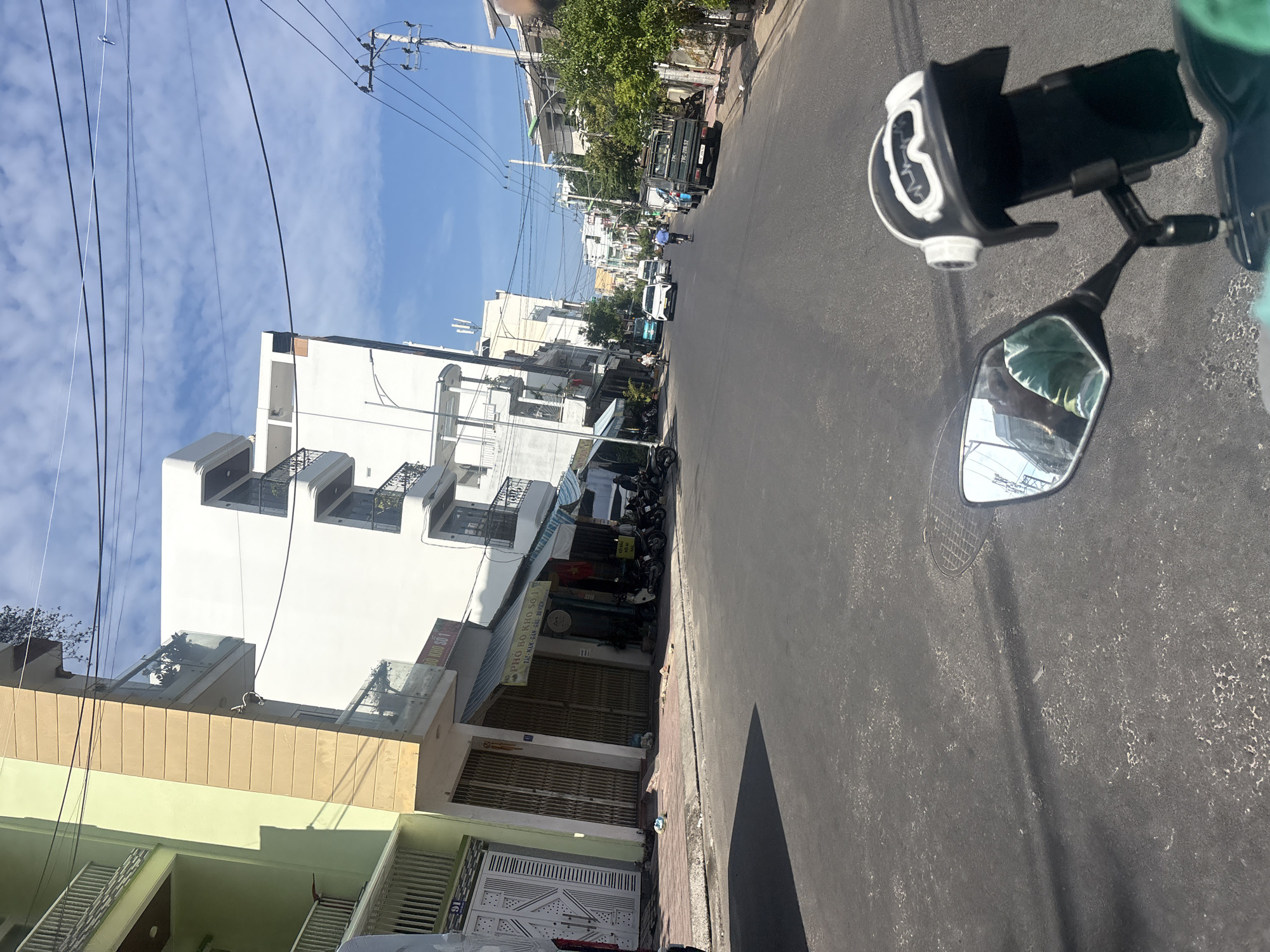 The image depicts a street scene in Nha Trang, Vietnam, likely taken from the perspective of a motorcycle rider. The street is paved with asphalt and appears to be in a moderately urbanized area. On the left side of the street, there are multi-story buildings with a light green facade on the left and a beige facade to its right. These buildings have balconies with railings and some potted plants. Further down the street on the left, there are shuttered storefronts. The right side of the street features more buildings, some with trees and greenery. Several parked motorcycles are visible along the sidewalk. In the middle of the street, there are cars and motorcycles moving, including a white car, a van, a blue motorcycle, and a dark green truck. The sky is blue with scattered white clouds. In the foreground, the mirror of a motorcycle shows a reflection of buildings and a person wearing a green shirt. A decorative helmet is mounted on a phone holder in front of the rider.