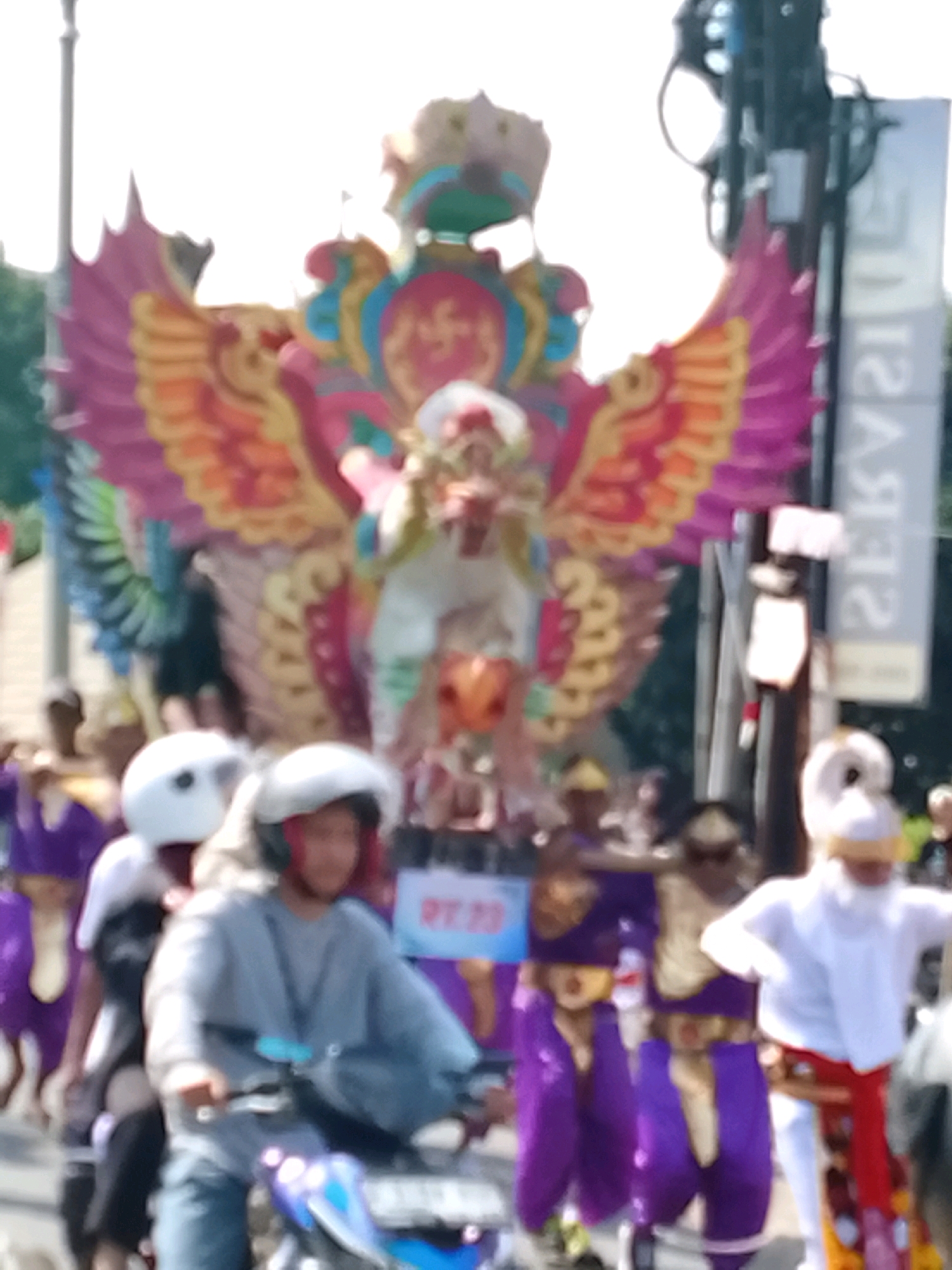 The image shows a blurred outdoor scene, possibly a parade or festival, in Ujungrusi, Indonesia. In the foreground, two individuals on motorcycles are partially visible, wearing helmets. The person on the right has red helmet accents and a grey top. In the background, a large, brightly colored effigy, likely representing a mythical creature with wings, is prominent. People in colorful costumes, some appearing to be purple and gold, are also visible in the procession. A tall vertical sign with white text on a grey background is to the right of the effigy. The overall lighting suggests it is daytime.