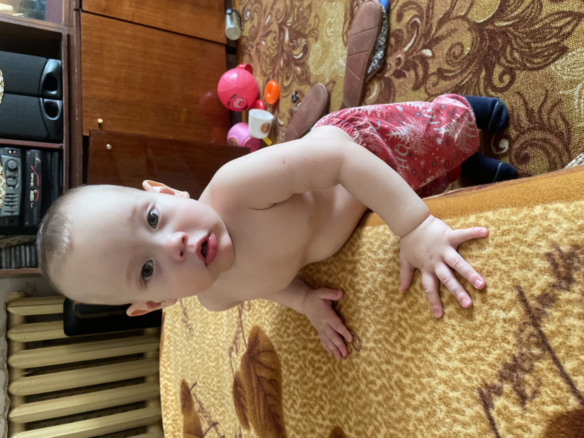 A baby lies on a patterned brown and tan blanket or bedspread. The baby is shirtless, wearing red patterned shorts and dark socks. The child is positioned near the edge of the surface, with their torso visible and their arms outstretched on the blanket.
To the left of the child are items on the edge of a wooden cabinet, including audio speakers and a stereo system. Behind the child are various items, including a pink ball, a cup, and slippers, on what appears to be an upholstered headboard. A radiator is partially visible on the left side.
The photo was taken indoors, possibly in Siva, Russia, based on the location provided.