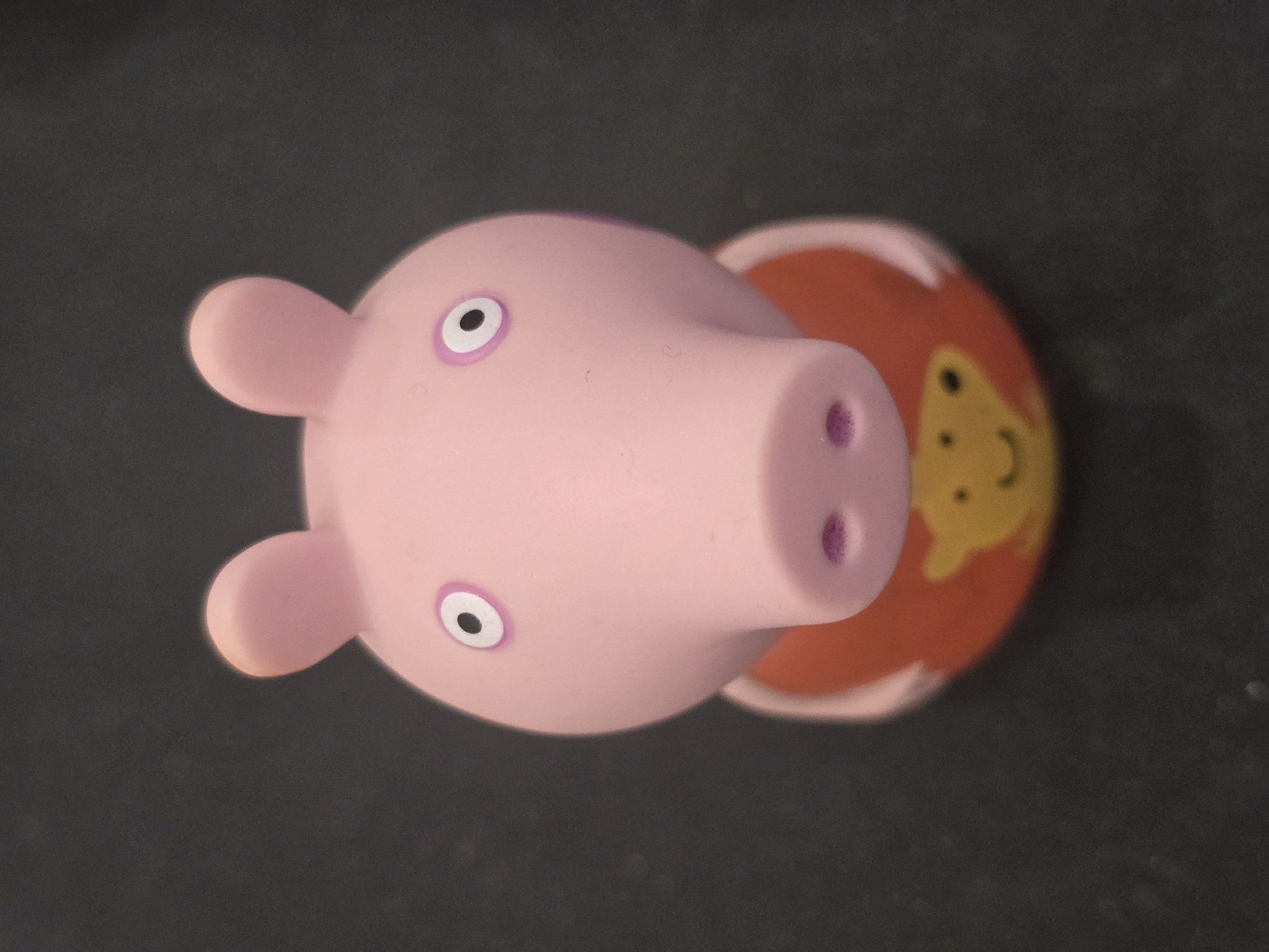 The image shows a close-up, overhead view of a pink rubber toy pig with large ears and a snout with two purple nostrils. It has two large, circular eyes with black pupils and pinkish-purple irises. The pig toy is placed on a dark, textured surface, possibly a table or floor. Behind the pig's head, a round, orange object with a yellow pattern is visible. The yellow pattern appears to be a simplified face with two eyes and a mouth. The overall impression is of a toy, possibly a bath toy, given its material and design. The lighting suggests it is indoors, with no indication of the time of day or weather. There is no visible text in the image.