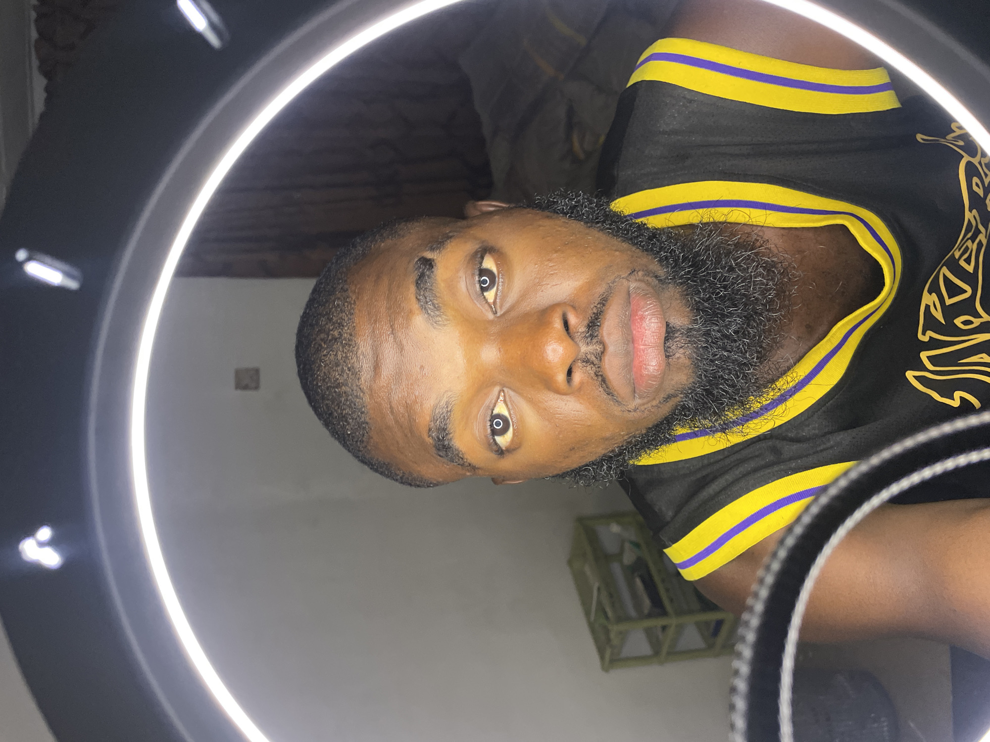 A close-up, medium shot captures a Black man with a beard and close-cropped hair, wearing a black basketball jersey with yellow and purple trim. He is positioned in front of a ring light, which casts a bright glow. His expression is neutral, and he looks directly at the camera. In the background, a small green shelf unit is visible, along with a blurred, dark wall and curtains. The overall setting appears to be an indoor space, possibly a bedroom or a studio, in Jos, Nigeria.