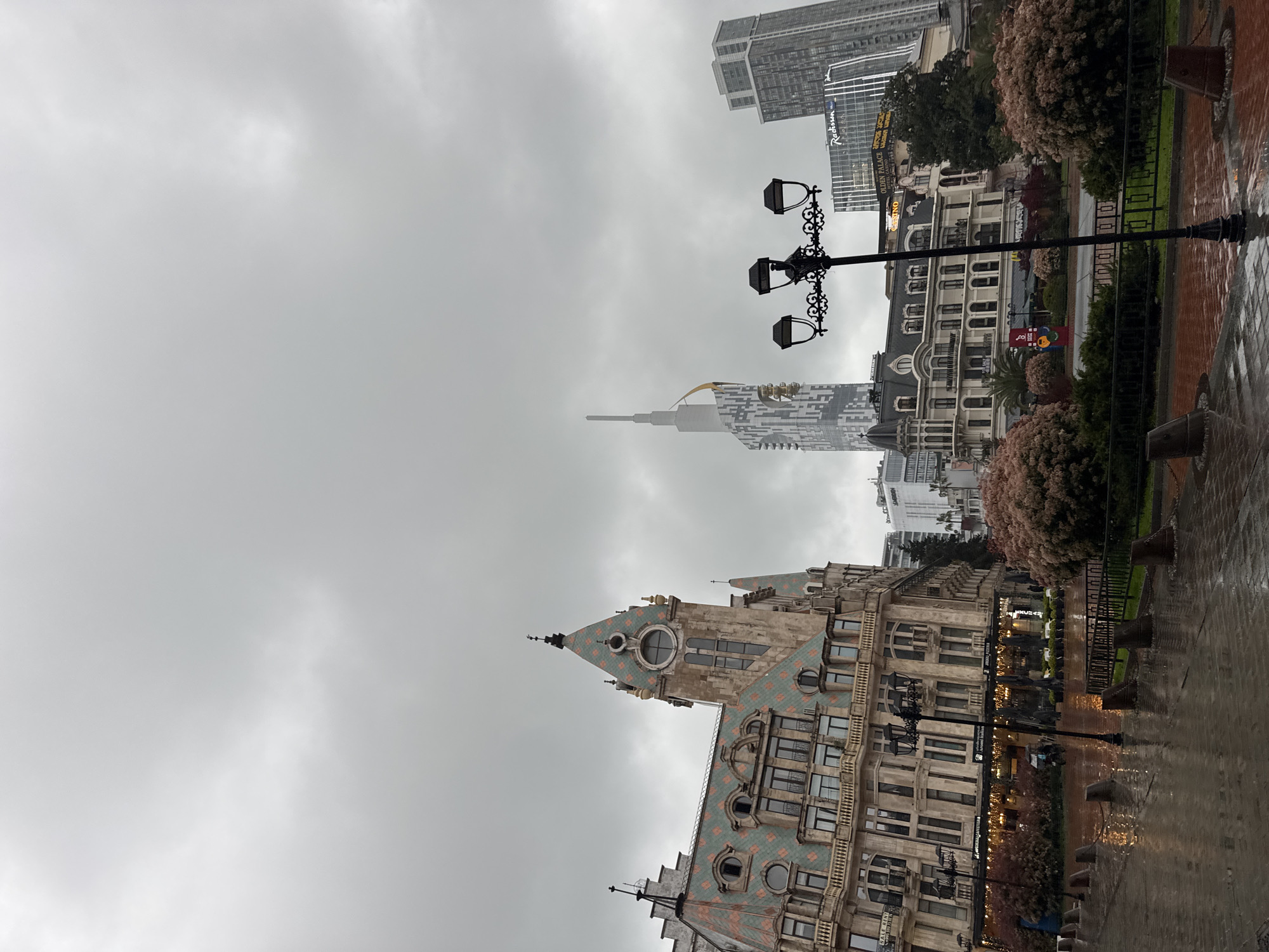 An urban street scene unfolds under an overcast, uniformly grey sky, indicating a cloudy or rainy day. The ground in the foreground and extending to the right is wet and reflective, suggesting recent or ongoing rain.
In the mid-ground on the left, a grand, ornate building with a distinctive peaked roof dominates. The roof is covered in a patterned mosaic of green and reddish-orange square tiles and features several round windows, one resembling a clock face near the peak. The building's facade is light-colored with numerous windows, some arched, and intricate architectural details.
To the right of this building, a black ornate street lamp post with three lanterns stands beside a black metal fence. Behind the fence and across the scene, there are lush bushes with light-colored blossoms or leaves. Further back, additional buildings are visible, including one with prominent signs for "QUEEN PALACE," "GOLDEN PALACE," and "CASINO," and another modern high-rise bearing the "Radisson" name. In the background, several tall, modern skyscrapers pierce the sky, one with a unique spire-like top and geometric patterns on its facade, and another more conventional rectangular tower.
There are no people visible, and no specific activity is taking place. The scene captures a mix of historical and contemporary architecture in a city environment during daytime, characterized by the muted light of a grey sky and wet surfaces.