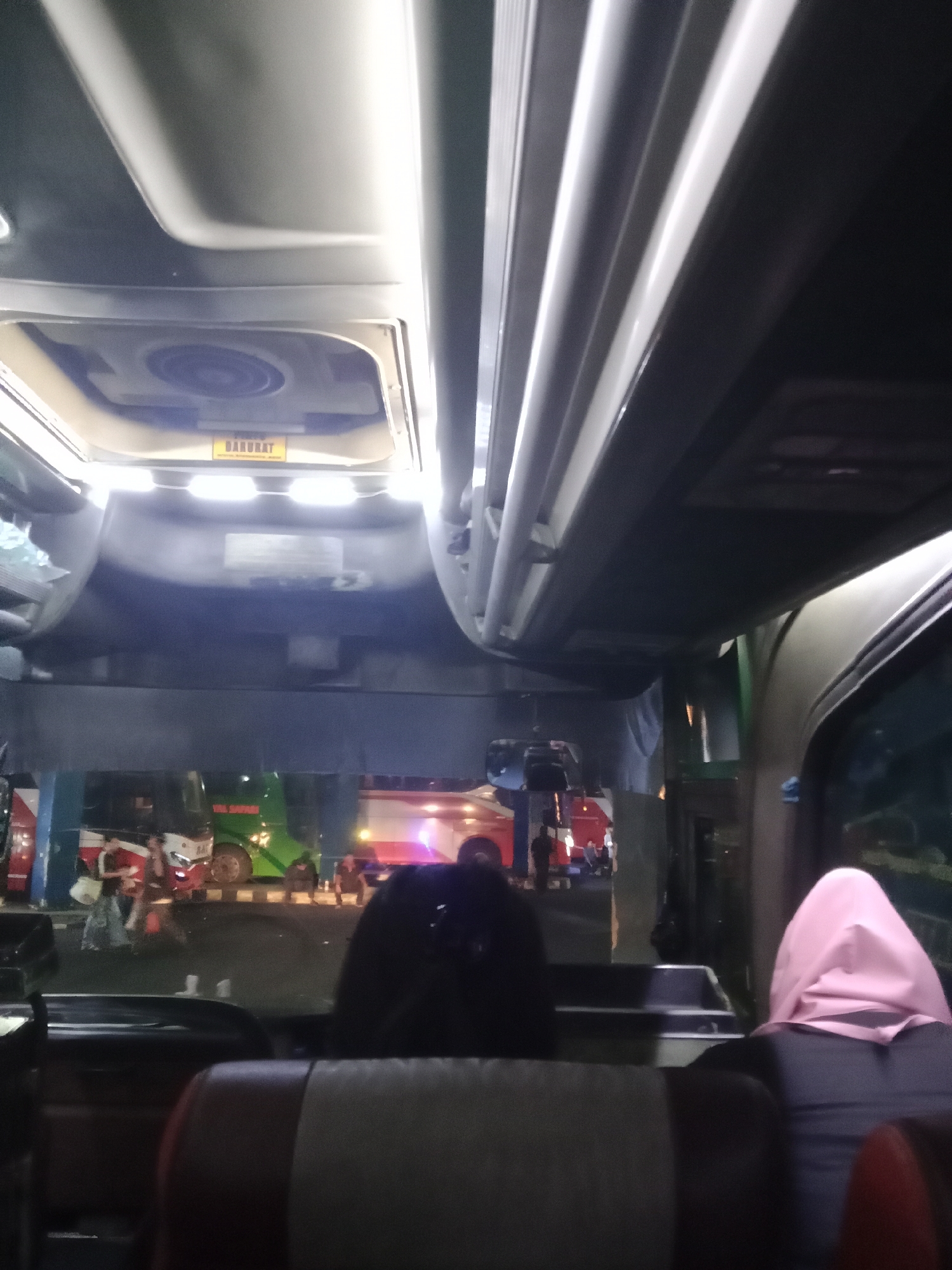 The image shows the interior of a bus, likely at a bus station in Surakarta, Indonesia. The perspective is from a passenger seat looking forward and slightly upwards. Visible are the bus's ceiling with overhead compartments and lighting. A yellow sign labeled "DARURAT" (Indonesian for "Emergency") is affixed to the ceiling, indicating an emergency exit or equipment. Through the front windshield, other buses and people are visible, suggesting a busy terminal. One bus in the background is green and white with "TULUS JAYA" written on it. A person wearing a pink hijab is seated in the foreground, facing away from the camera. The overall lighting is dim, with bright highlights from the bus's interior lights.