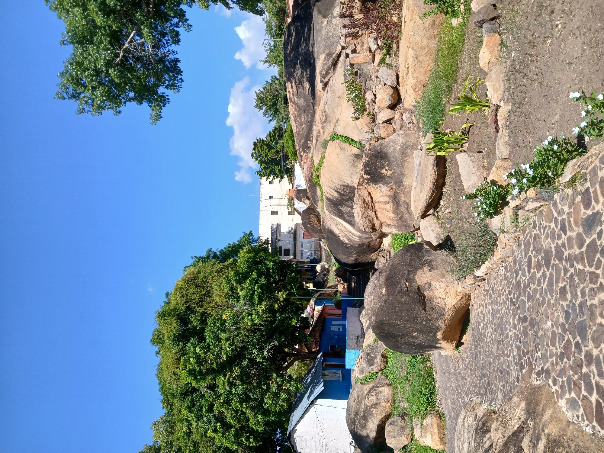 The image shows a scene with rocks, vegetation, and houses. A mosaic-patterned walkway is visible. There is a blue and white building partially obscured by a large tree. The sky is blue with some clouds. A multi-story building is visible in the background.