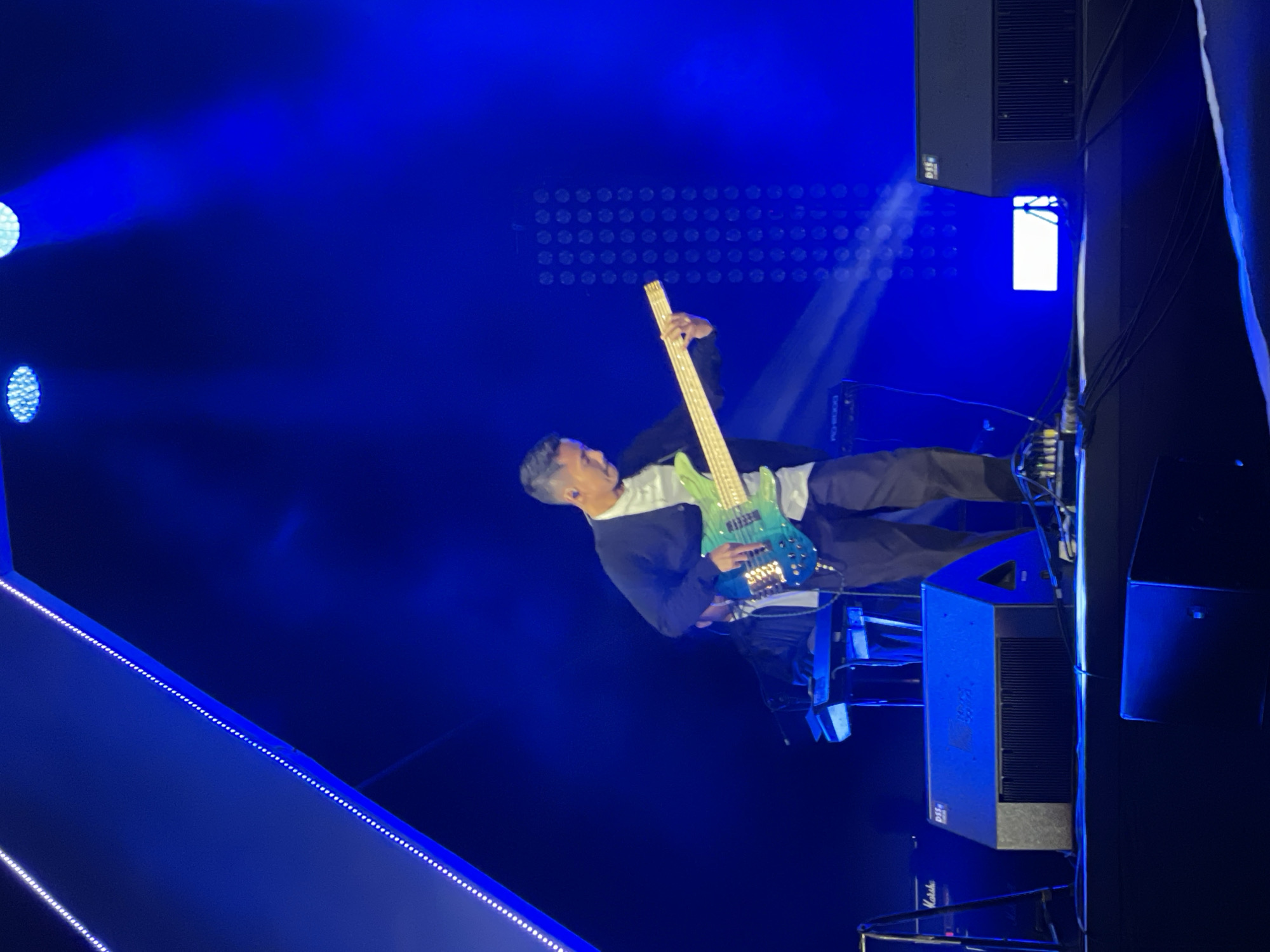 A man is playing a bass guitar under blue stage lighting. The location is a stage in Salembaran, Indonesia. The man is wearing a dark jacket over a light-colored shirt and dark pants. The guitar has a light-colored neck and a blue body. Speakers and other stage equipment are visible. A strip of LED lights is partially visible on the left side.