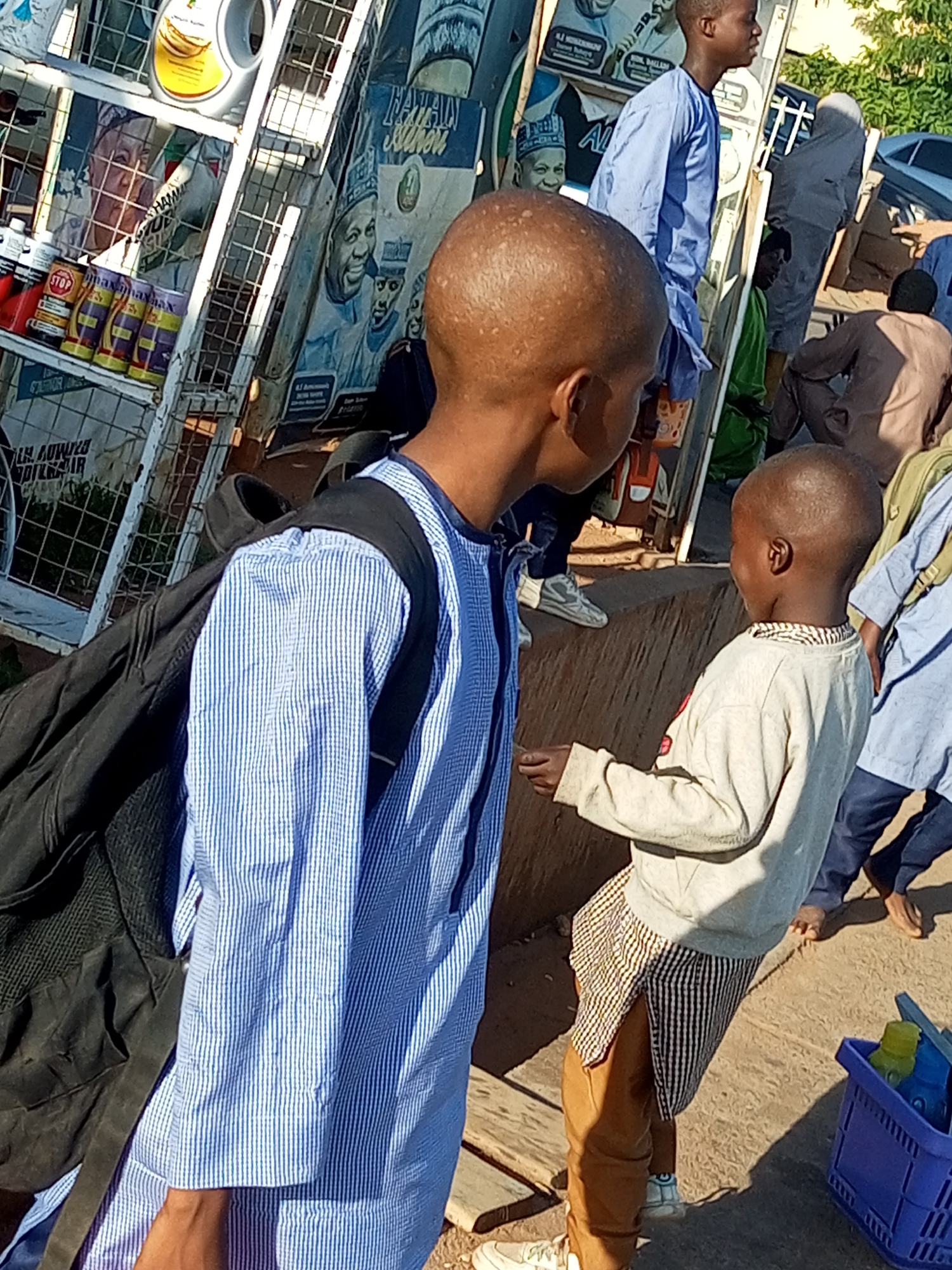 In Gombe, Nigeria, a group of young boys are gathered around a market stall displaying various goods. In the foreground, a boy with a shaved head wears a blue and white striped shirt and carries a black backpack. To his right, another boy in a light-colored long-sleeved shirt and patterned shorts stands with his hand extended. Behind them, a market stall features shelves stocked with bottled drinks, cleaning products, and posters of political figures. Several other individuals, including adults, are visible in the background, some engaged in conversation or observing the surroundings. The scene suggests a bustling outdoor marketplace environment.