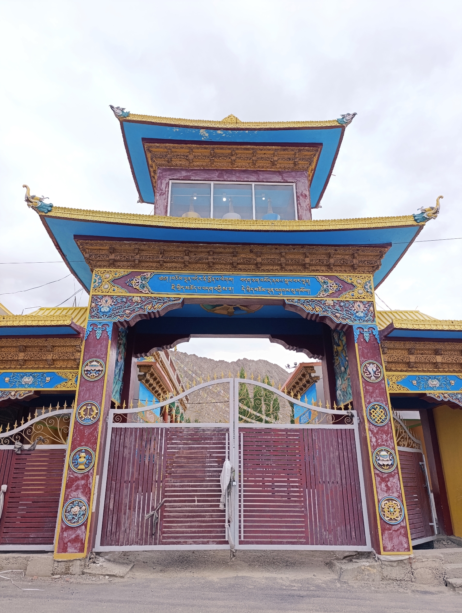 A low-angle, full shot captures the ornate entrance gate of a Buddhist monastery in Leh, India, under a cloudy sky. The gate features a multi-tiered, pagoda-style roof adorned with gold trim and blue accents. Decorative dragon figures with yellow accents perch on the edges of the upper roofs. The central archway is framed by colorful, intricate carvings and Tibetan script in blue. Below the archway, large maroon gates, made of vertical wooden planks, are partially open. To the sides of the main gate are decorative metal railings. The overall structure exhibits vibrant colors, including blues, golds, reds, and purples, typical of Buddhist architectural styles. The background reveals a glimpse of a mountainous landscape.