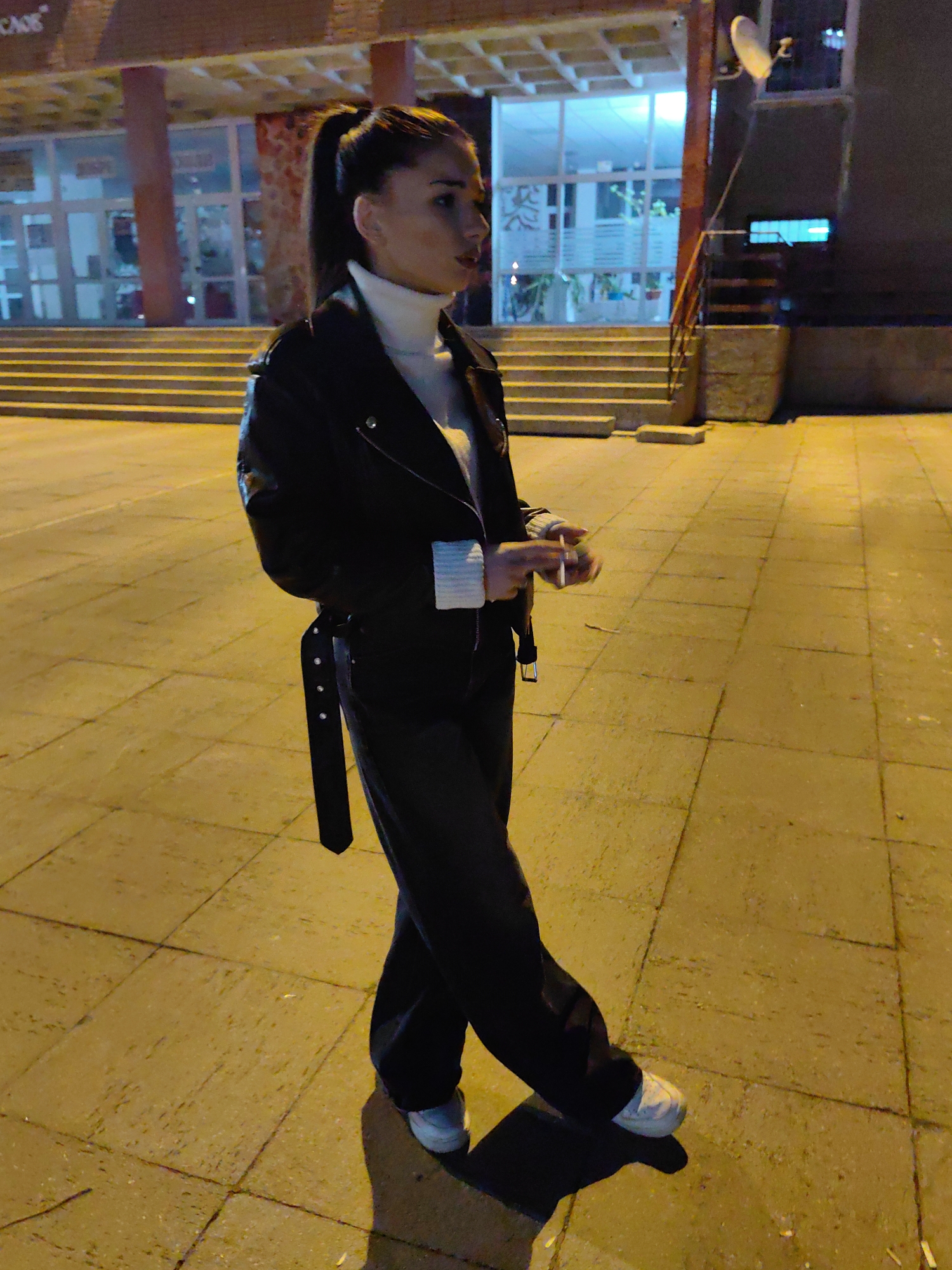 A female individual stands on tiled pavement outdoors at night, observed in a partial left profile. She wears a white turtleneck sweater, a black leather jacket, dark trousers, and white athletic shoes. A black belt with silver-toned embellishments hangs from her left hip. In her right hand, she holds a lit cigarette. Her gaze is directed towards the right side of the frame.
The ground consists of light-colored rectangular pavement tiles, illuminated by warm yellowish-orange artificial light which creates distinct shadows. In the background, to the left and center, is a building with a large glass facade entrance. A wide set of concrete steps leads up to this entrance, flanked by reddish-brown support columns. Partial text reading "CADE" is visible at the top left of the building. The interior, visible through the glass, is lit with cool blue-white light and shows vegetation. To the right, a darker building section features a white satellite dish mounted high on its facade and a horizontal blue light fixture. This architectural style, particularly the expansive glass entrance and grand staircase, is characteristic of structures located in the vicinity of the National Palace of Culture (NDK) in Sofia, Bulgaria.