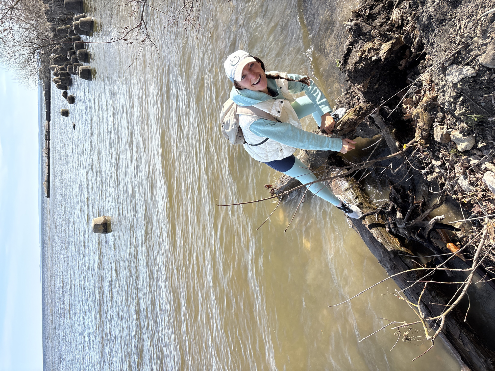 A young woman is captured smiling cheerfully at the muddy bank of a wide river, likely in Ulyanovsk, Russia, on a bright, sunny day. The image is taken at a diagonal angle, making the horizon appear tilted.
The woman is positioned at the water's edge, appearing to interact with or climb upon exposed tree roots and a large log that extend from the bank into the murky, greenish-brown water. She is dressed in a light blue long-sleeved top and leggings, an off-white quilted vest, and a light-colored baseball cap. A light-colored backpack is slung over her shoulders. Her dark hair is styled in a braid, and her expression is one of genuine happiness.
The river water shows distinct ripples, reflecting the clear blue sky overhead. In the distance, along what appears to be the opposite bank or a structure extending into the water, numerous dark, square or rectangular concrete blocks or structures are visible, partially submerged. Bare tree branches, characteristic of late winter or early spring, hang over the water from the immediate riverbank. The bank itself consists of dark, damp soil, intertwined roots, and scattered dry leaves and debris. There is no visible text on her clothing or the surrounding environment. The scene conveys a sense of outdoor adventure and enjoyment of nature.