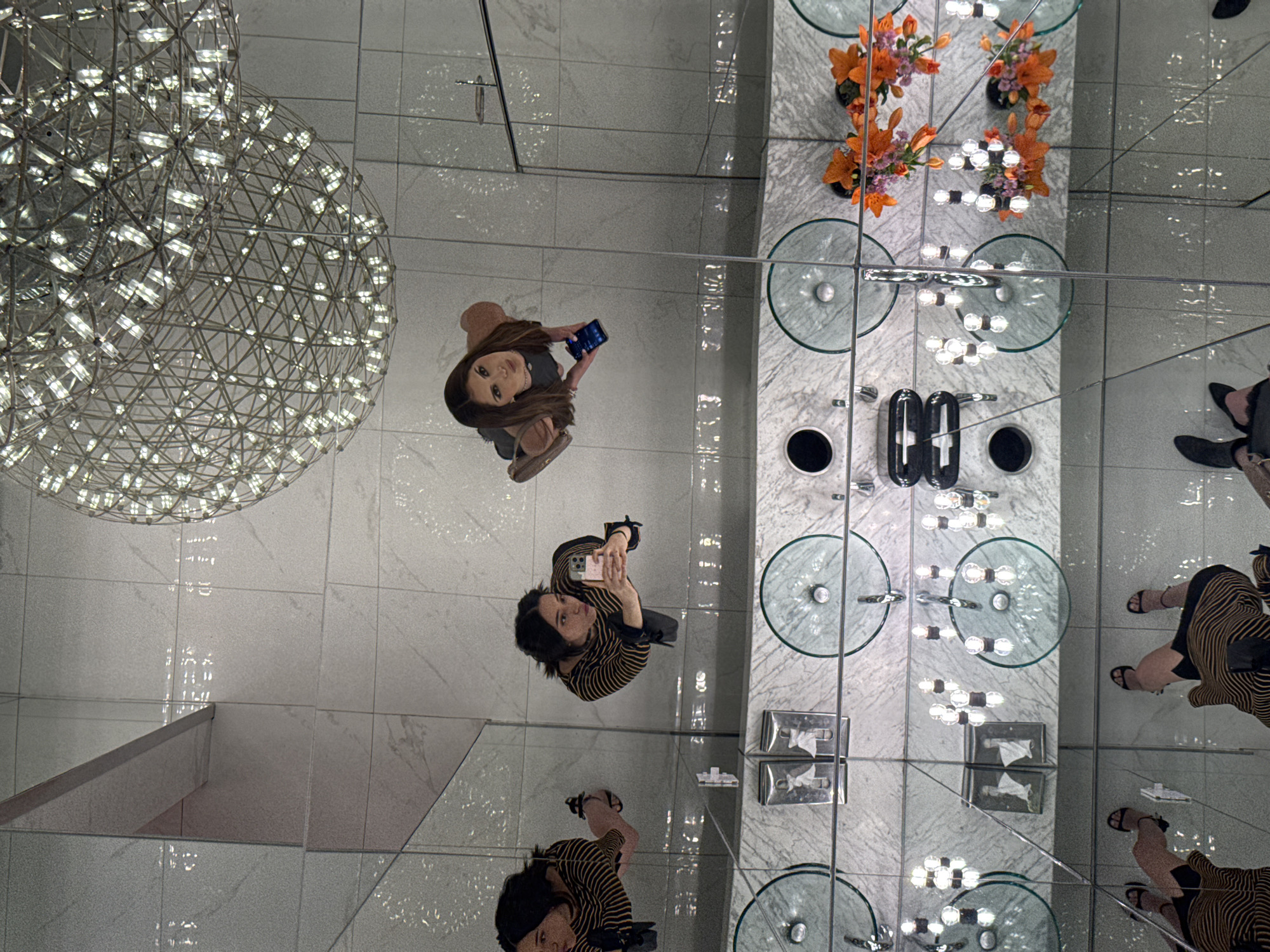A top-down perspective photograph depicts the interior of a room with mirrored surfaces. The room appears to be a bathroom with multiple sinks, a light fixture that resembles a sphere made of intersecting metal lines with small lights. Three people are visible. One person is standing near the sinks, holding a mobile phone. The second person is near the sinks, holding a mobile phone. The third person is located near the sinks. The sinks are glass with metallic fixtures. Oranges and flowers are placed on the sinks. There is tiling on the wall and floor. The location may be in Puebla, Mexico.