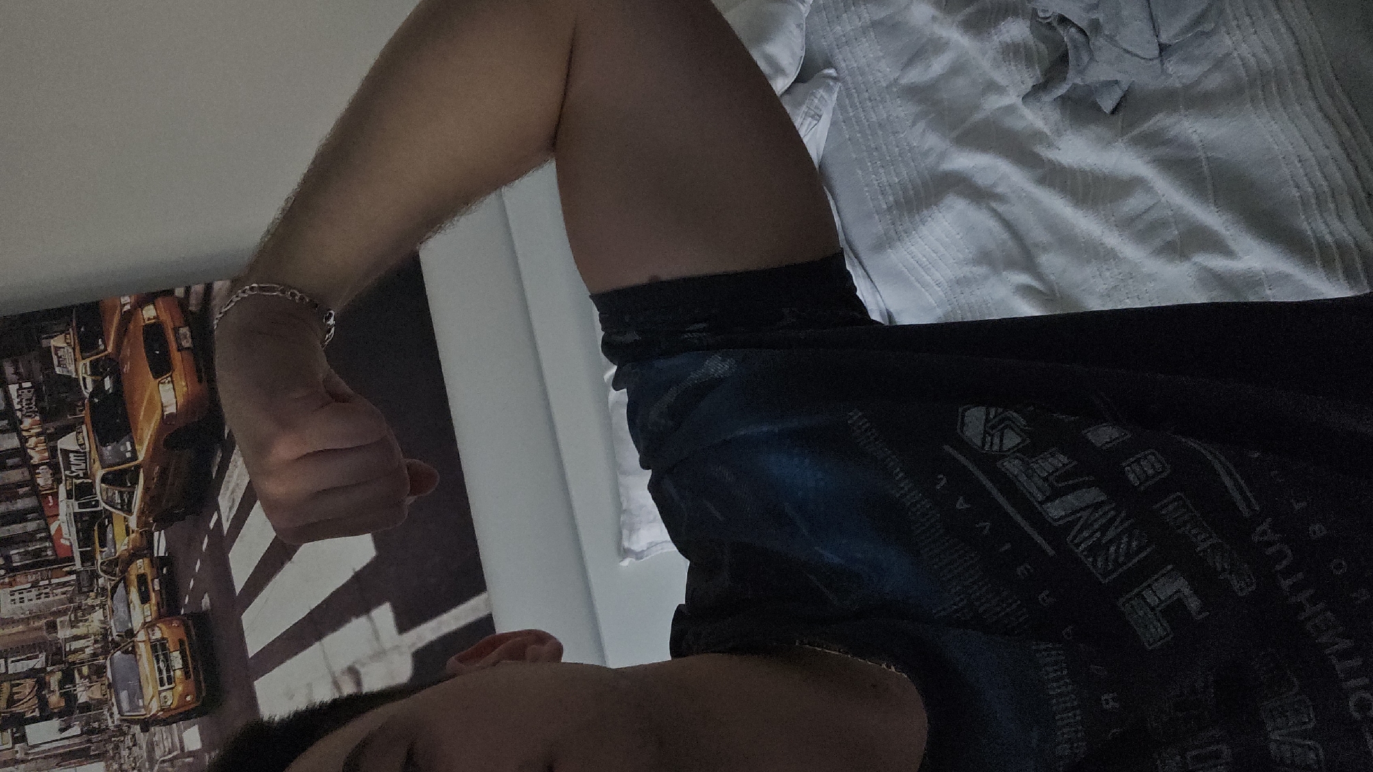 A male person is positioned with their head towards the lower portion of the frame, and their torso extends diagonally upwards, towards the right. The person is wearing a dark t-shirt with printed text and designs. The person's left arm is flexed upwards, forming an "L" shape, and their fist is clenched. The arm is adorned with a silver bracelet. The backdrop behind the arm and hand features a poster or framed image of a cityscape, with several yellow taxi cabs depicted. Above the person, there is a white textured bedding or fabric. The location in the city of Plovdiv, Bulgaria is not discernible in the image.