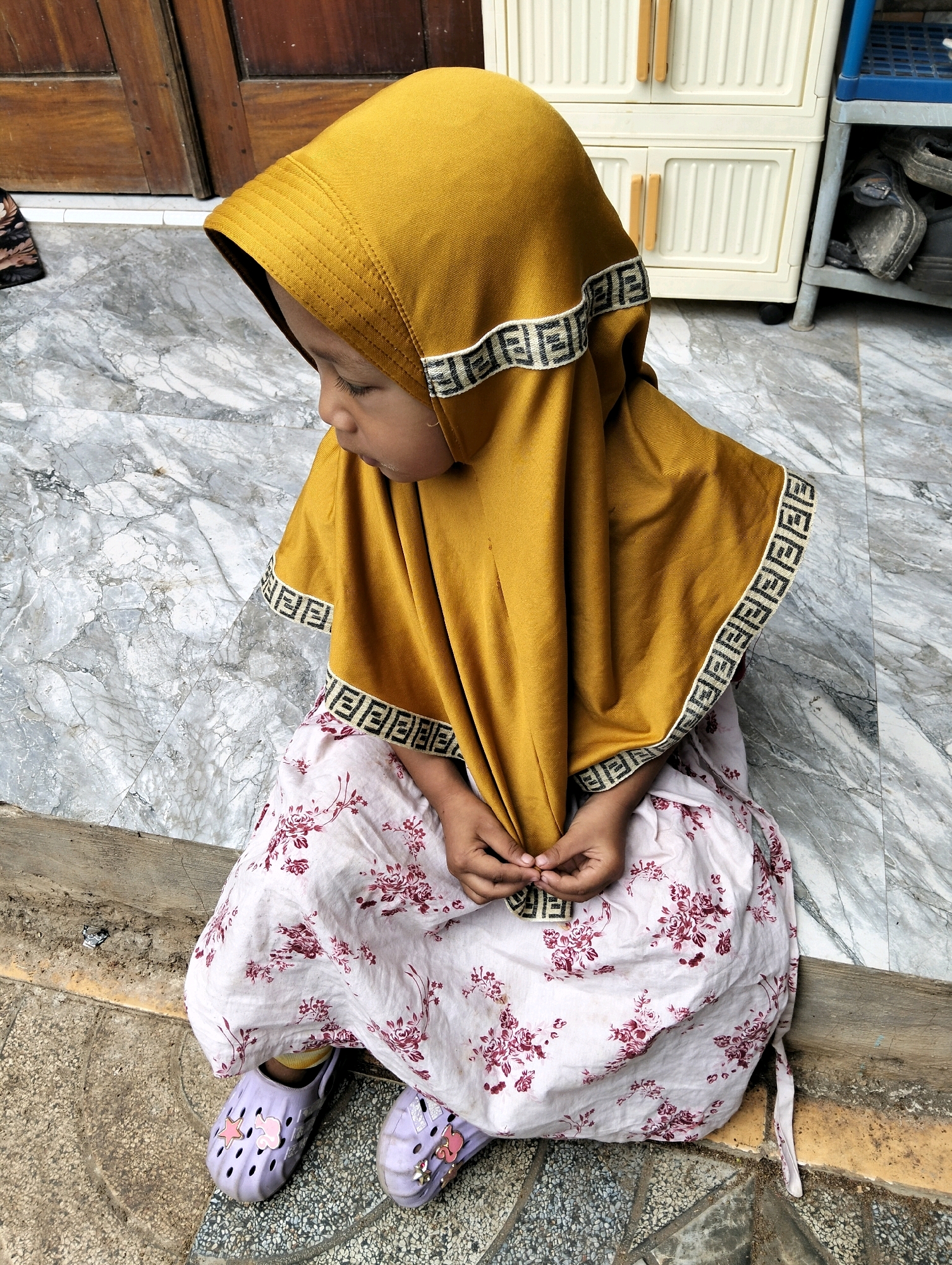 In Cileungsi, Indonesia, a young child, likely female based on clothing, sits on a marble-patterned tiled floor with a wooden door visible in the background. The child wears a mustard-yellow hijab with a black and white decorative trim and a light pink dress with a floral pattern. She is also wearing light purple clogs decorated with pink charms, including a star. Her hands are clasped in her lap. To the right of the child, a cream-colored plastic cabinet with two doors is visible, and further to the right, a blue shelving unit contains dark-colored shoes. The overall scene suggests an outdoor or semi-outdoor setting, possibly a porch or walkway.