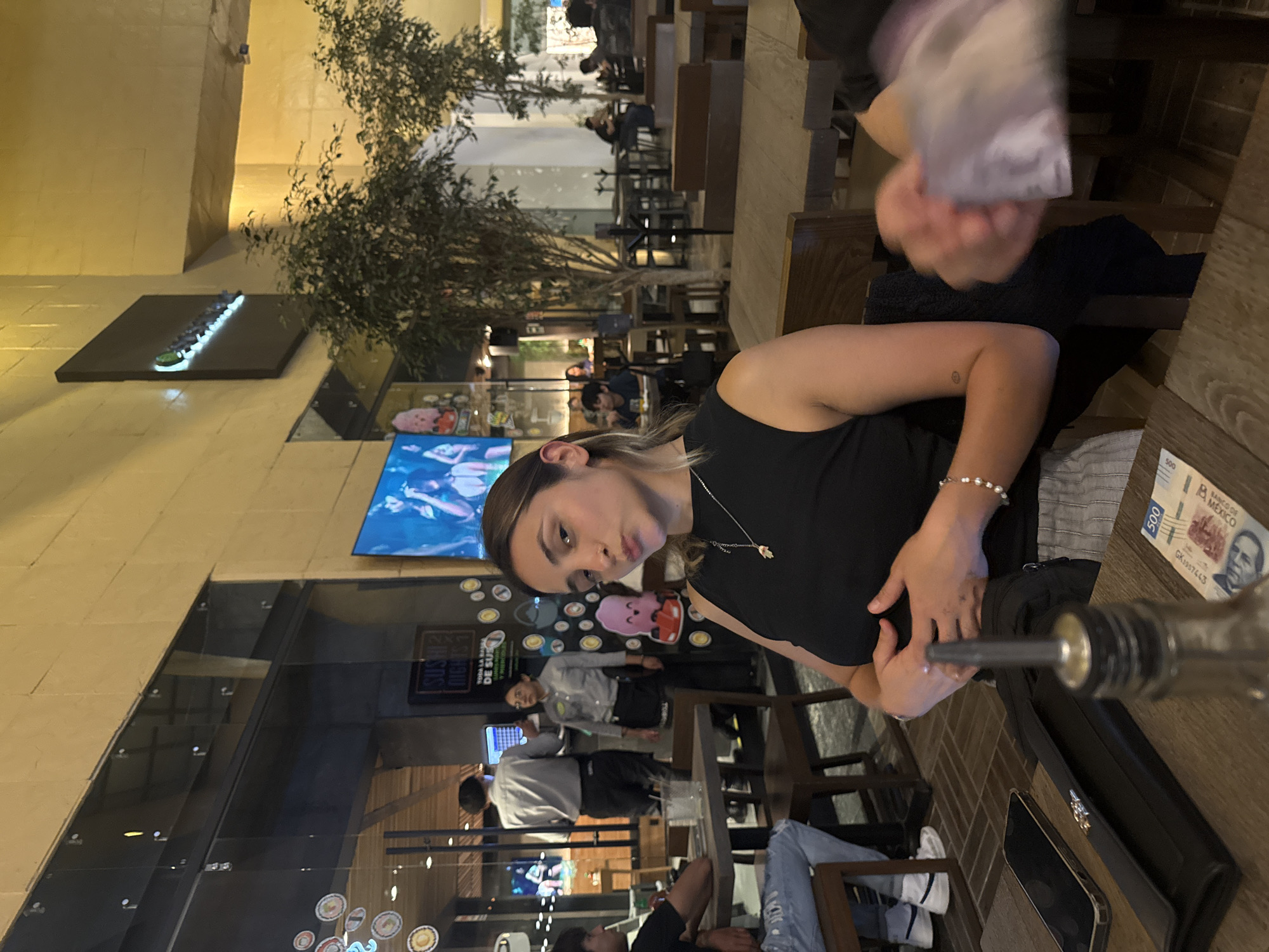 A woman with blonde hair is sitting at a restaurant table. She wears a black tank top. The setting appears to be in Puebla, Mexico, with a Mexican 500-peso note visible on the table. A black purse is also visible on the table. The restaurant seems to have an outdoor seating area with trees and a view of a street or plaza. A waiter or other staff member is standing in the background near a glass partition.