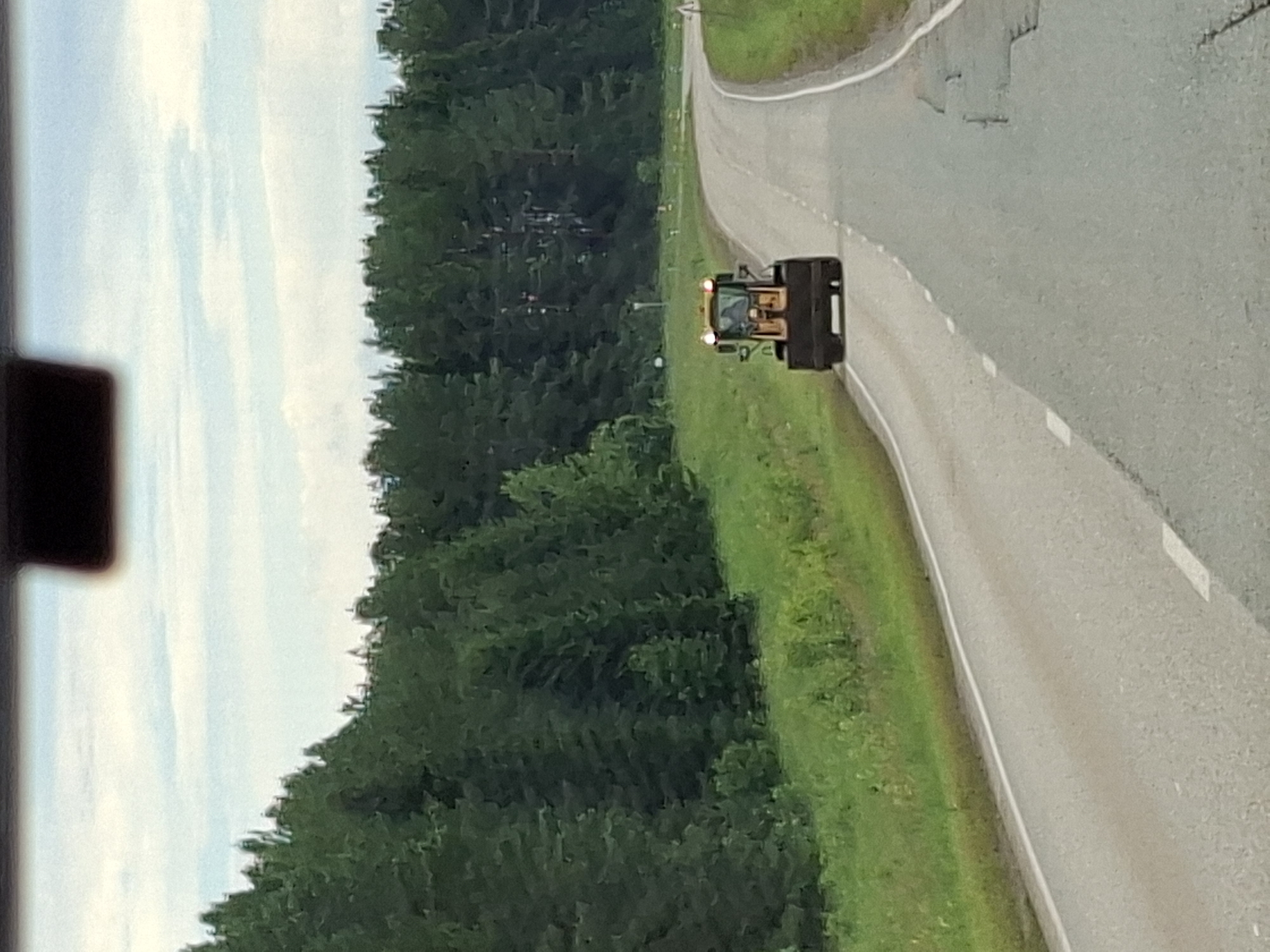 This media file depicts a yellow front-end loader driving on a paved road, with its bucket lowered. The loader has visible headlights and what appears to be a driver inside the cab. The scene is set in a rural or semi-rural environment. To the left of the road, there is a dense forest of green trees. To the right of the road, there is a grassy verge. The sky above is cloudy and light. The image is taken from a slightly elevated perspective, possibly from a vehicle. The overall impression is of a daytime scene, likely during fair weather, given the visibility. The setting appears to be outdoors, with the road curving gently to the right. No text is visible in the image. The location context is Ляльшур, Russia.