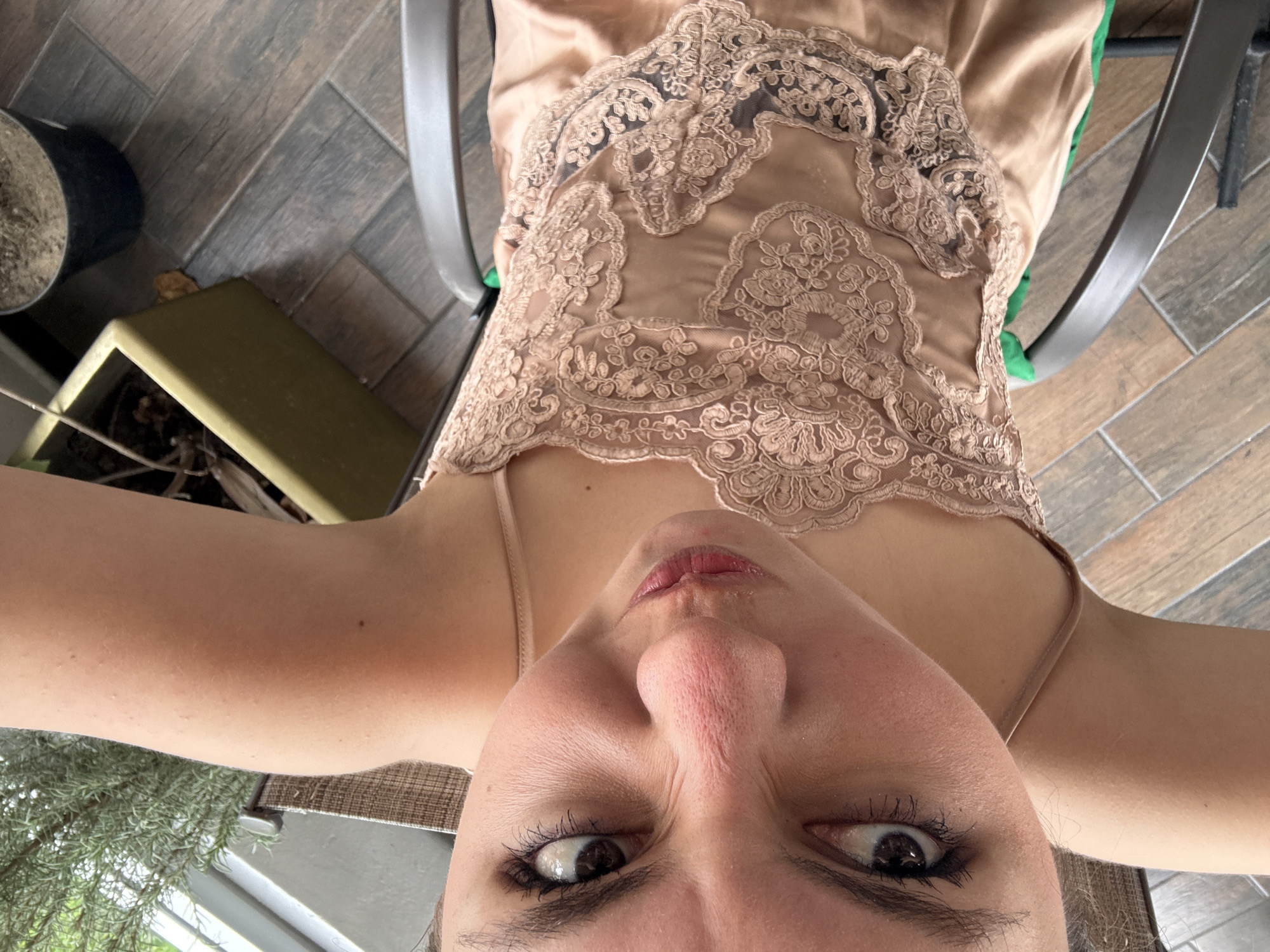 The image presents a close-up, downward-angled view of a woman's face and upper torso. She is wearing a light brown, lace-trimmed top with spaghetti straps. Her face is characterized by a prominent nose, pursed lips, and visible makeup around the eyes. The background reveals a patio or outdoor area with grey-toned tiled flooring and potted plants. There is a green, boxy plant pot and a black pot, with a window visible in the lower left corner. The chair is a grey metal frame. There is no information that suggests the photo was taken in Puebla, Mexico.