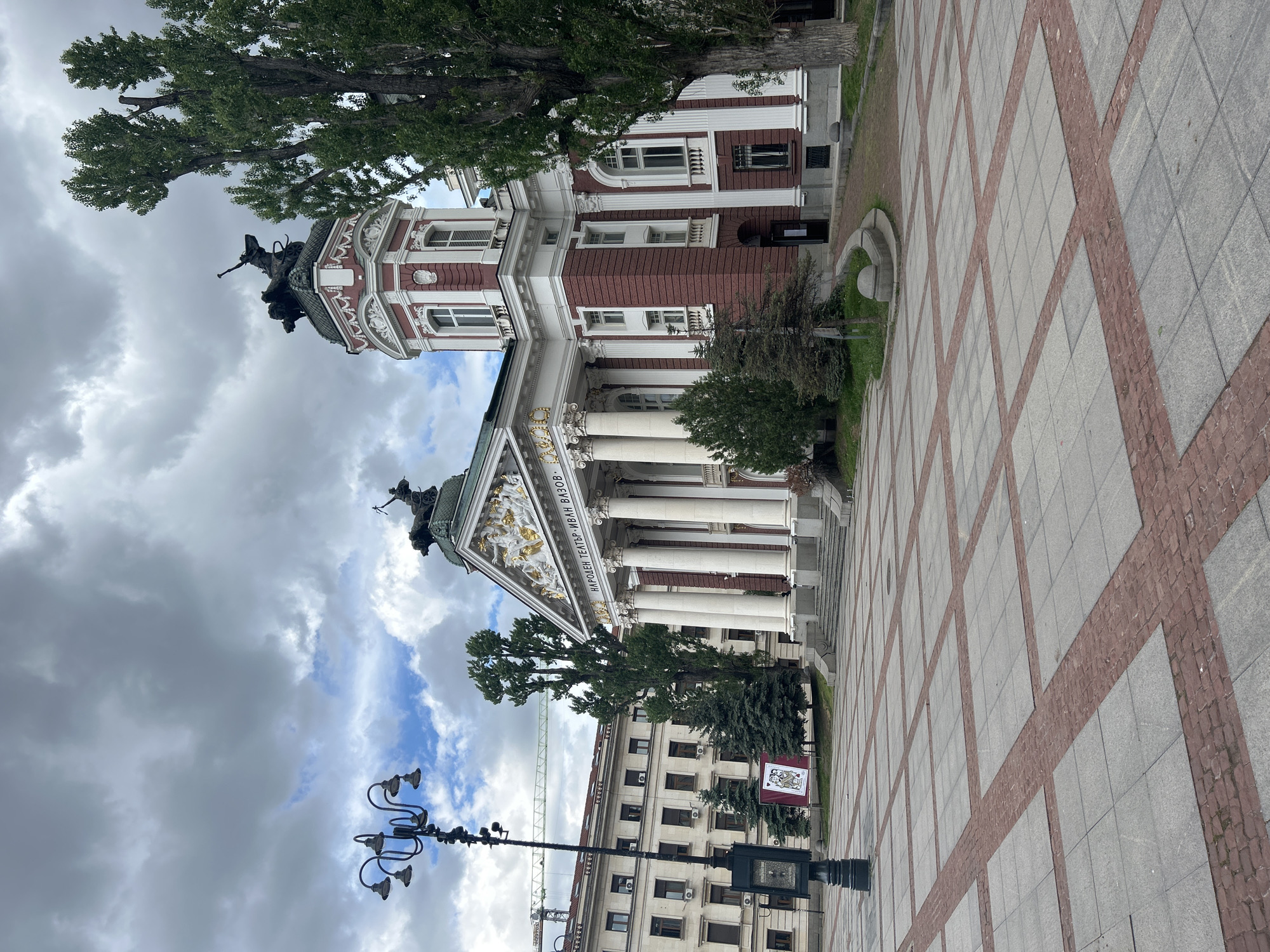 The image shows the Ivan Vazov National Theatre in Sofia, Bulgaria. The theatre is a grand building with a classical facade, featuring columns and statues. The sky above is cloudy. A lamppost is visible in the foreground. The ground is paved with gray stones and red bricks.