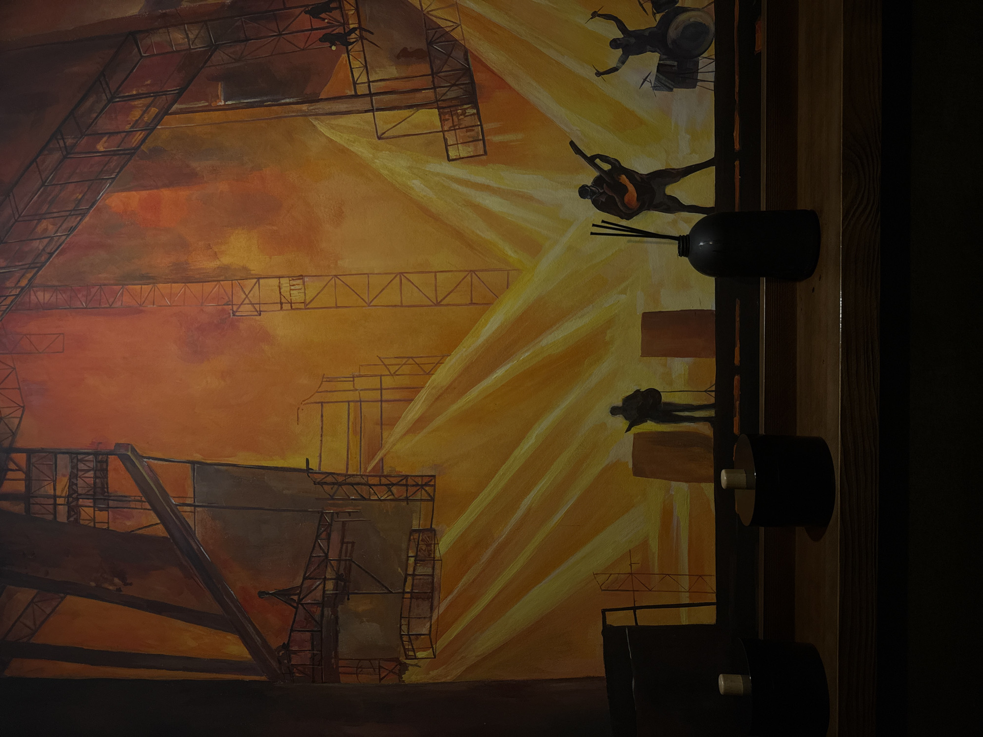 This image captures a wide shot of a large, vibrant mural depicting a concert stage or industrial setup, located in Izhevsk, Russia. The mural features a warm color palette dominated by rich oranges and yellows, suggesting intense stage lighting.
The upper portion of the mural illustrates a complex network of dark, structural scaffolding and trusses against a fiery orange background, with several small, silhouetted human figures positioned on different levels, appearing to be workers or stagehands. Intense yellow beams of light diagonally traverse the middle and lower sections of the painting, emanating from unseen sources above, creating a dynamic visual effect.
In the lower half of the mural, silhouetted figures of a band are depicted on a stage. From left to right, one figure stands next to two brown, barrel-like objects, holding what appears to be a guitar. Another figure in the center plays an electric guitar, with a microphone stand in front of them. To the far right, a third figure is shown vigorously playing a drum set, with drumsticks raised. The figures are rendered in dark silhouettes, emphasized by the strong backlighting.
In the immediate foreground, a dark wooden shelf spans horizontally across the bottom of the image. Three distinct objects are placed on this shelf: two dark, cylindrical containers with light-colored, possibly wooden, lids or stoppers are positioned on the left side. To the right, a dark, matte black bottle containing several thin, dark reeds, consistent with a reed diffuser, stands prominently. The environment beyond the mural and shelf is dark, indicating an indoor setting with subdued ambient lighting, allowing the mural to be the focal point.
