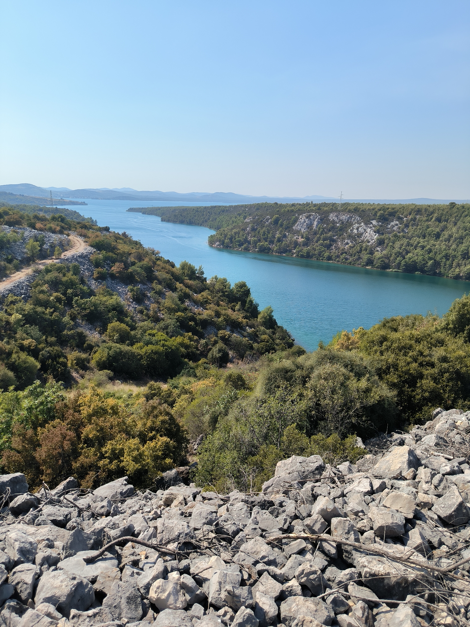 This image depicts a scenic view of a wide, tranquil river surrounded by lush, green vegetation and rocky terrain. The river, which appears to be a part of the Krka River, flows through a canyon-like landscape, with steep, tree-covered banks on either side. The foreground is dominated by a pile of large, grey rocks, some of which are covered in dry twigs and branches, suggesting a natural, perhaps slightly wild, environment.
In the middle ground, the river stretches out, its surface a striking shade of turquoise blue under a clear, bright sky. The riverbanks are densely forested with a variety of trees, their leaves a mix of vibrant green and some autumnal hues of yellow and brown. A winding dirt path is visible on the left bank, curving up the side of the hill.
In the background, rolling hills and distant mountains are visible, fading into a hazy horizon. The overall impression is one of natural beauty and a serene, untouched landscape. This scene is consistent with the natural beauty found within the Krka National Park, located in the city of Šibenik, Croatia, a region renowned for its stunning river system and Mediterranean flora. There are no people or man-made structures prominently featured in the image, emphasizing the natural state of the environment.