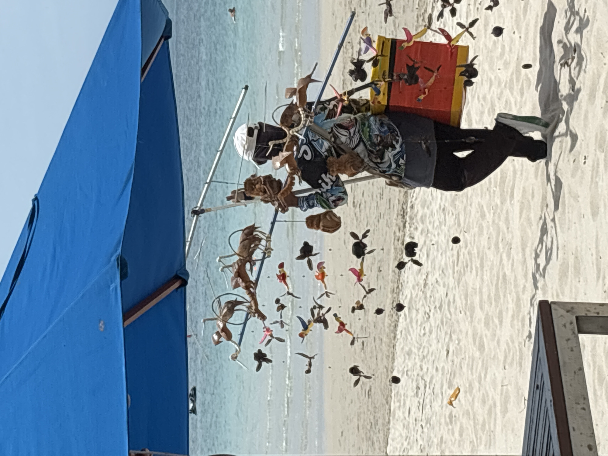 A vendor stands on the sandy beach of Baan Chaweng, Thailand, under a large blue umbrella. The vendor, wearing a white hat, face covering, and a colorful patterned shirt, is carrying a mobile display of wooden toys on a stand. The toys, predominantly in the shape of birds and animals, are suspended from poles and wires, some appearing to be in motion due to the wind. Behind the vendor is the turquoise ocean with gentle waves and the horizon under a clear sky. In the foreground, a portion of a dark slatted table is visible, suggesting a seating area. The beach is composed of light-colored sand.