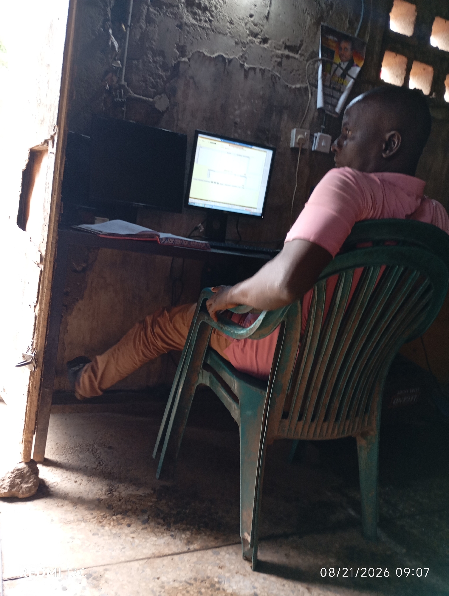 The image depicts a man sitting in a plastic chair in front of a desk with two computer monitors. The desk is made of wood and appears to be in a rustic setting, possibly an outdoor or semi-outdoor workspace. The walls are rough and unfinished, and there's a framed picture of a person on the wall. The floor is concrete. The man is wearing a pink shirt and brown pants. The scene is lit by natural light coming from the left and artificial light from a bulb, creating shadows. A date and time stamp "08/21/2026 09:07" is visible in the bottom right corner, suggesting the photo was taken on this date and time. The location is identified as Shendam, Nigeria.